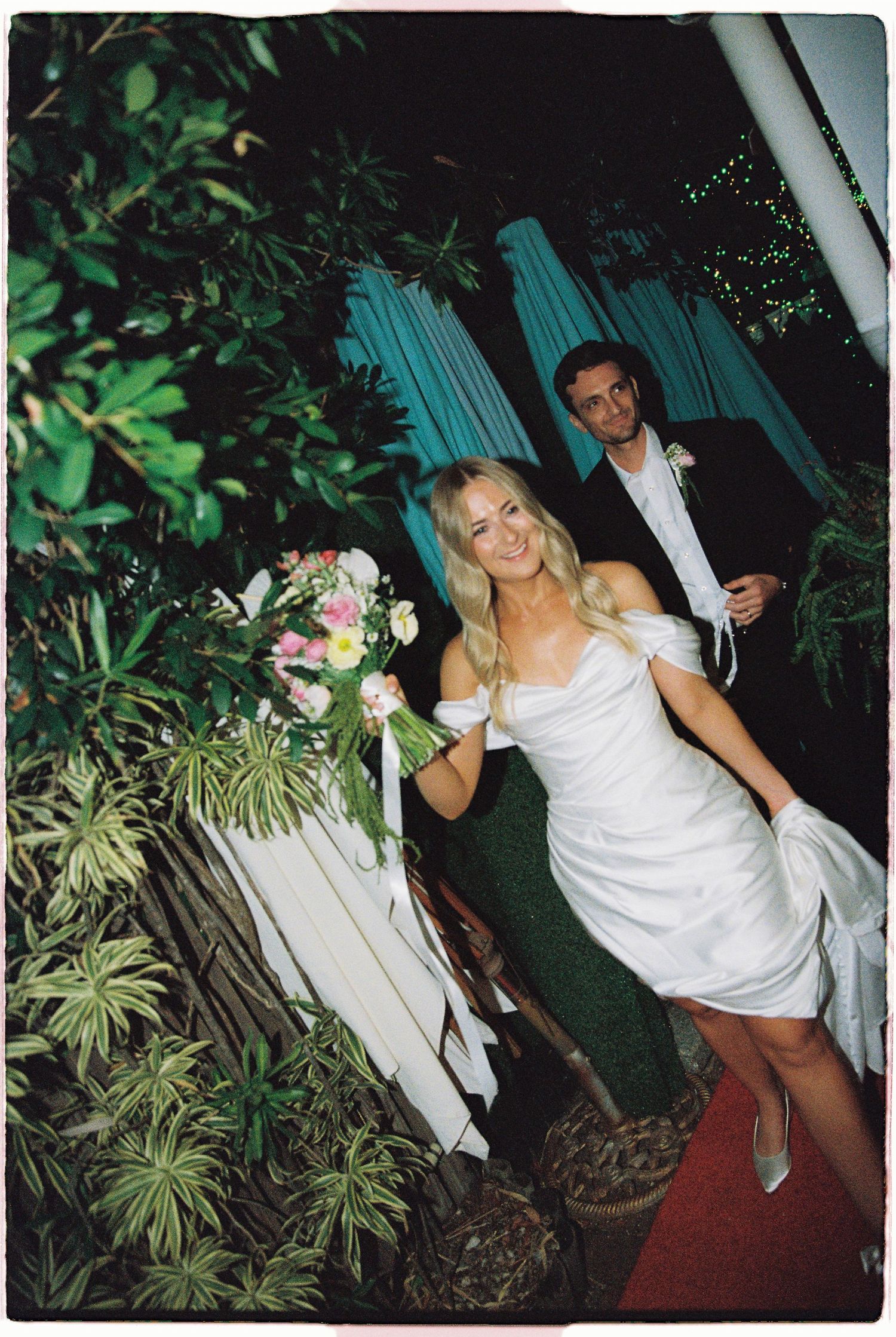 Bride in short white off-shoulder dress holding floral bouquet walks with groom on red carpet outdoors.