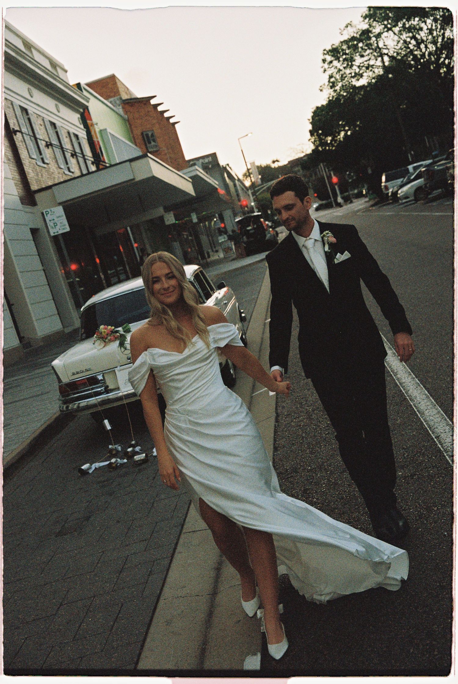 Bride in white off-shoulder gown and groom in black tuxedo walking hand-in-hand on a city street.