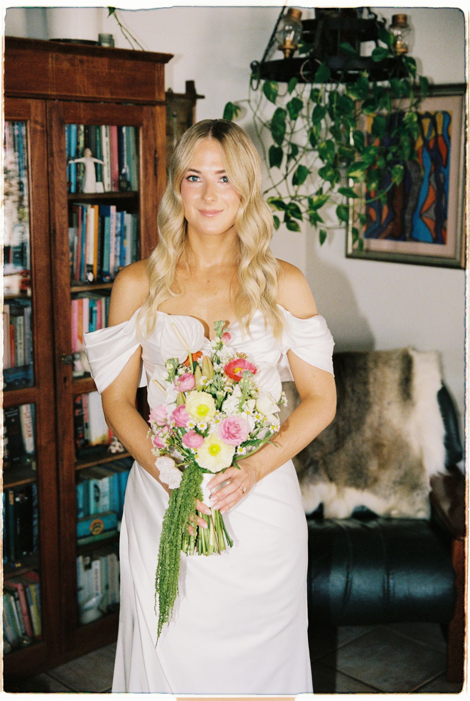 Blonde bride in off-shoulder white dress holding colorful spring bouquet indoors.