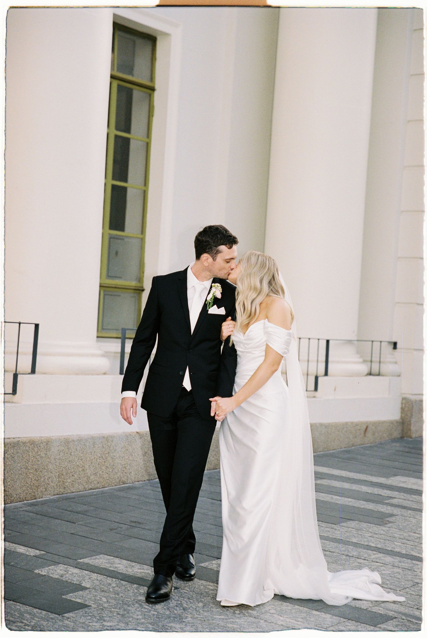 Bride and groom share a tender kiss on the steps of a grand white columned building.