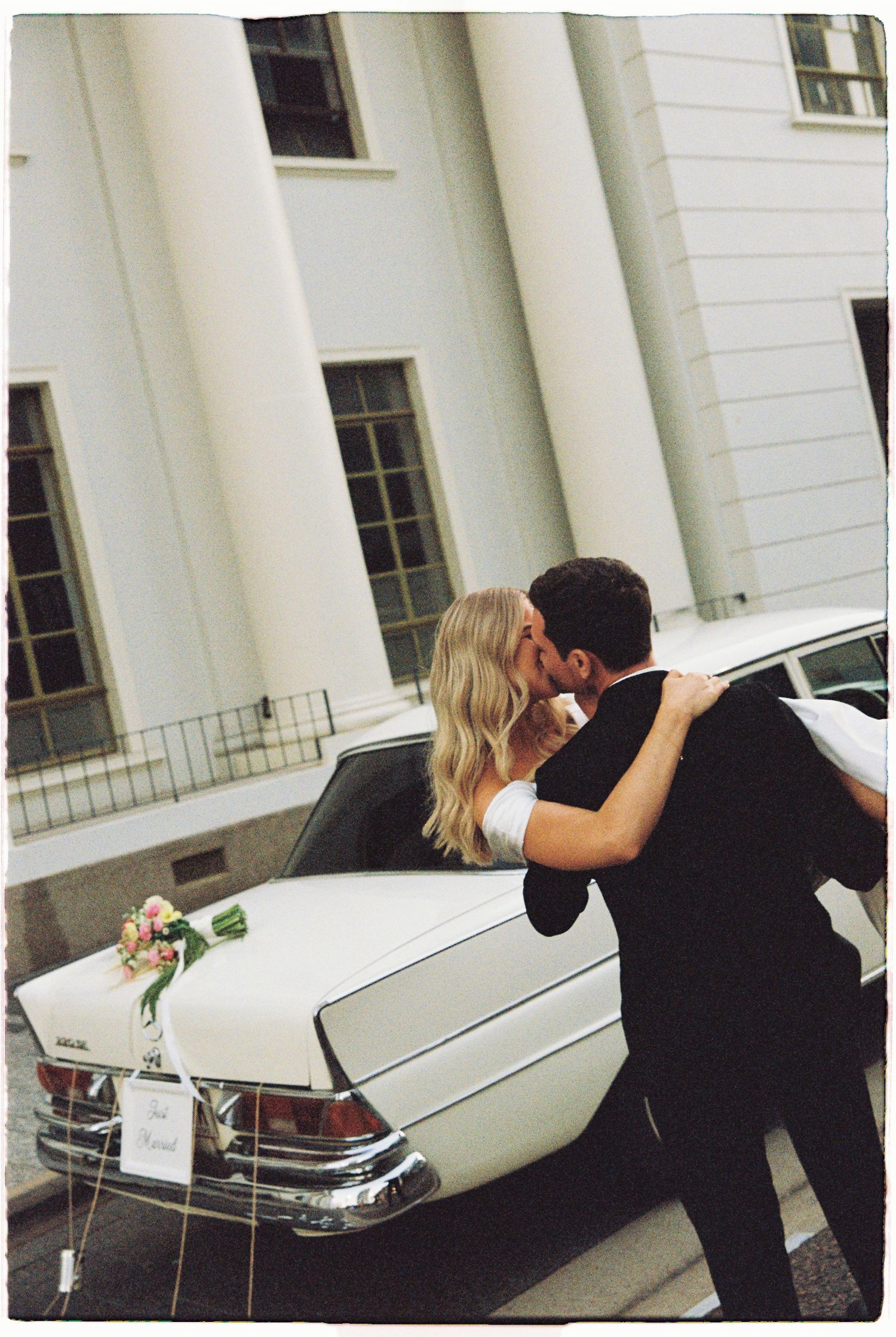Newlyweds share a kiss leaning against a decorated vintage white car outside a grand white building.
