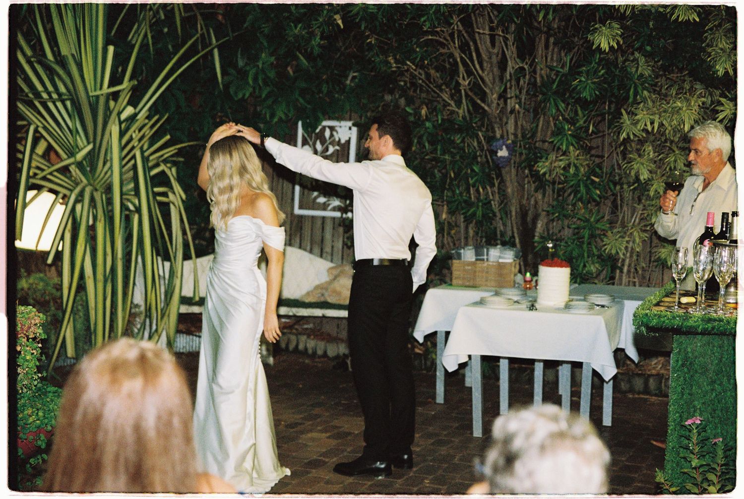 A groom in a white shirt twirls his blonde bride in a white dress on an outdoor patio wedding dance floor.