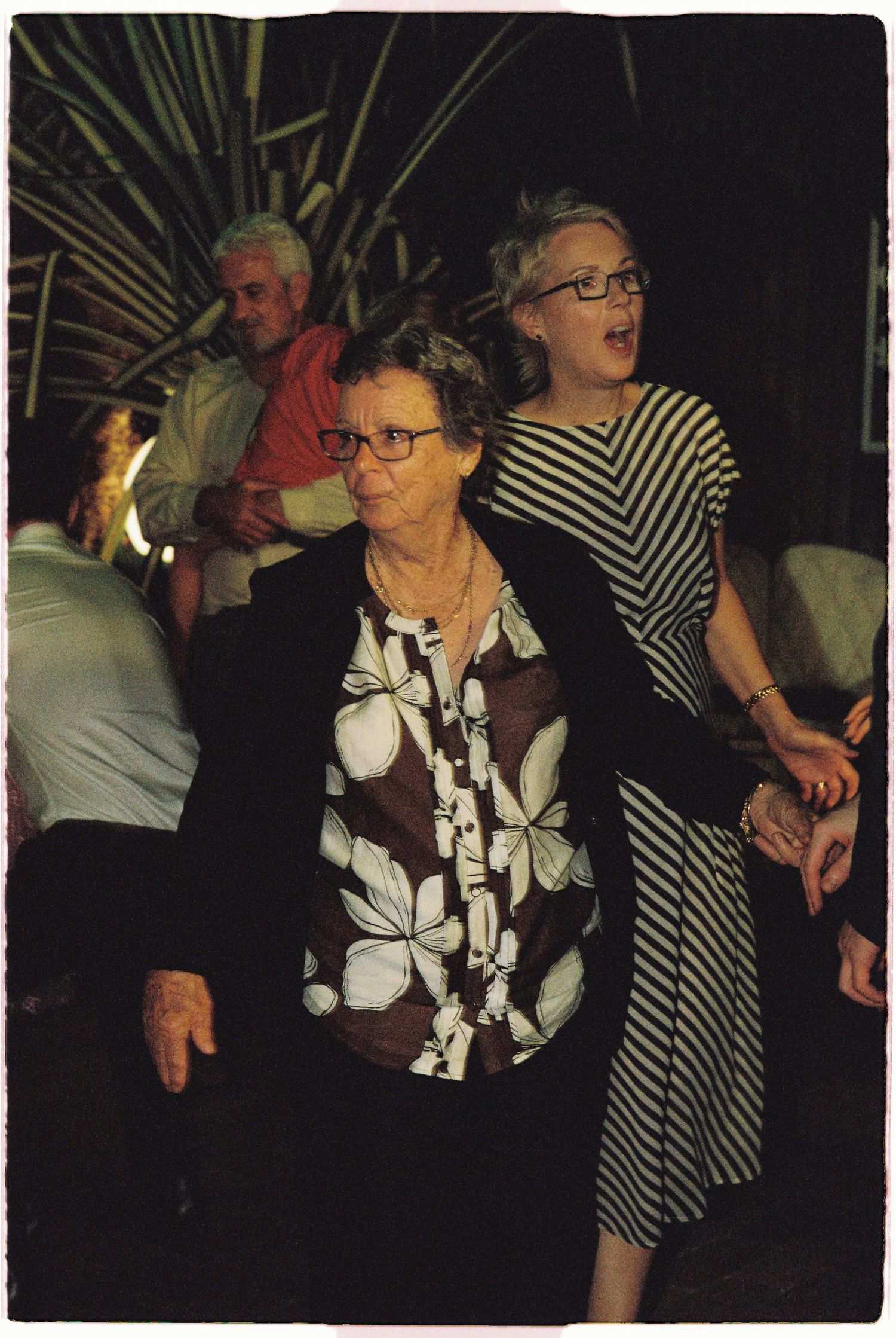 Two women dancing at evening wedding reception, one in floral blouse and blazer, other in striped dress with glasses.