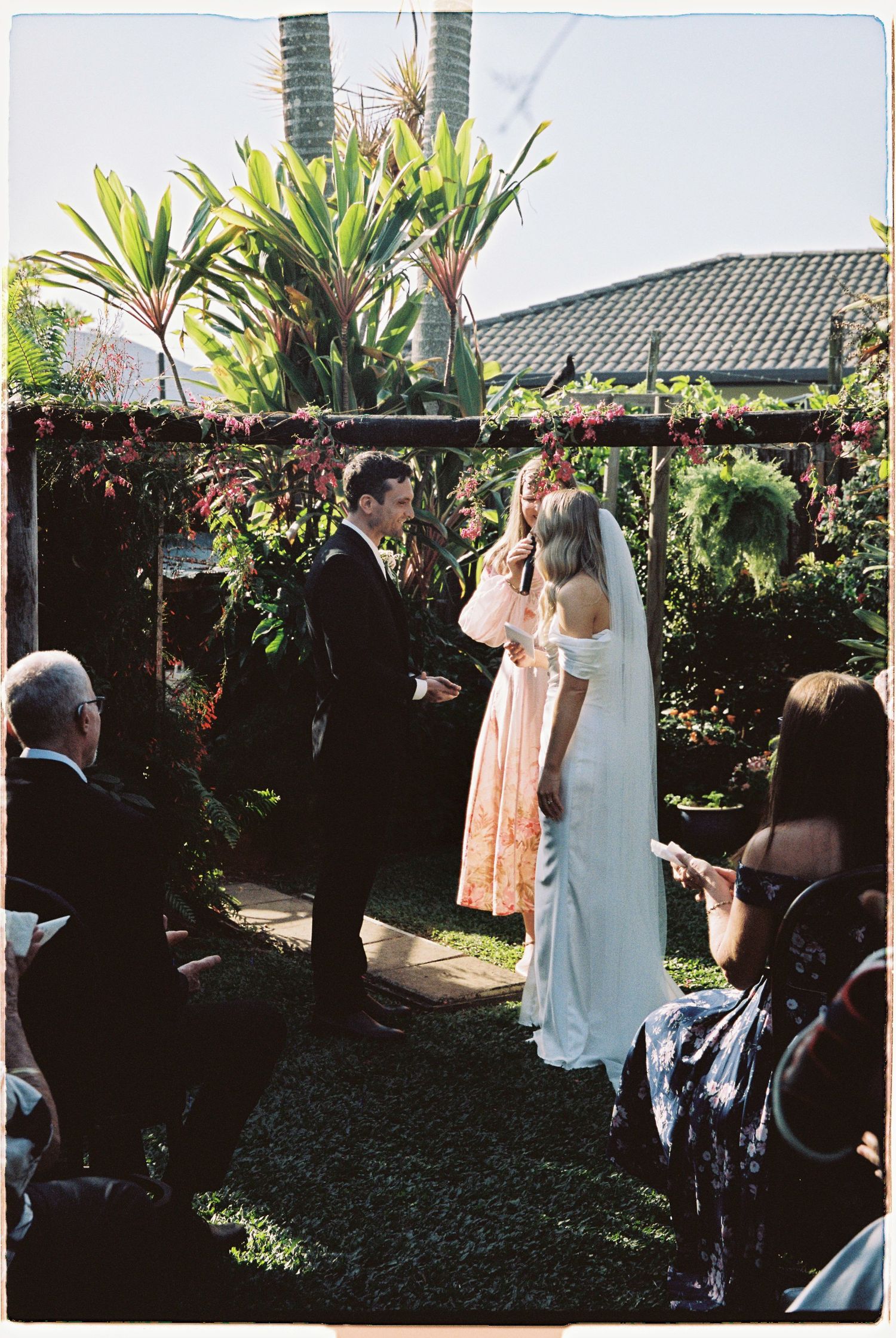 Bride and groom exchange vows under floral arch in tropical garden ceremony.