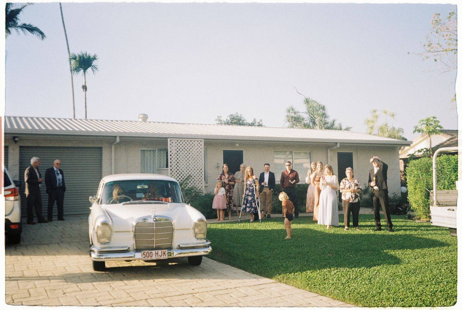 Guests gathered outside suburban home beside vintage white Mercedes wedding car on sunny day.