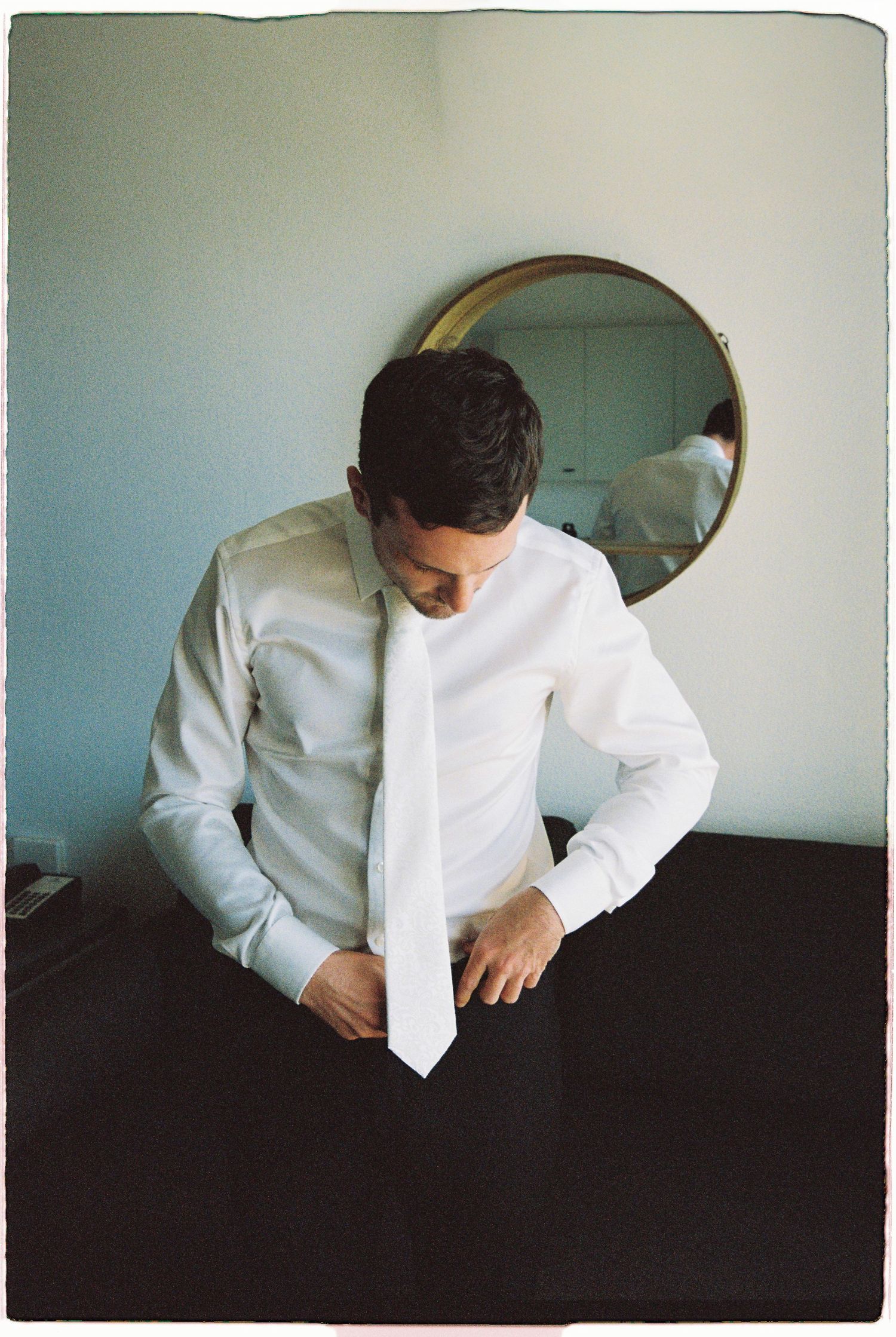Groom in white shirt adjusting white tie before mirror during wedding preparation.
