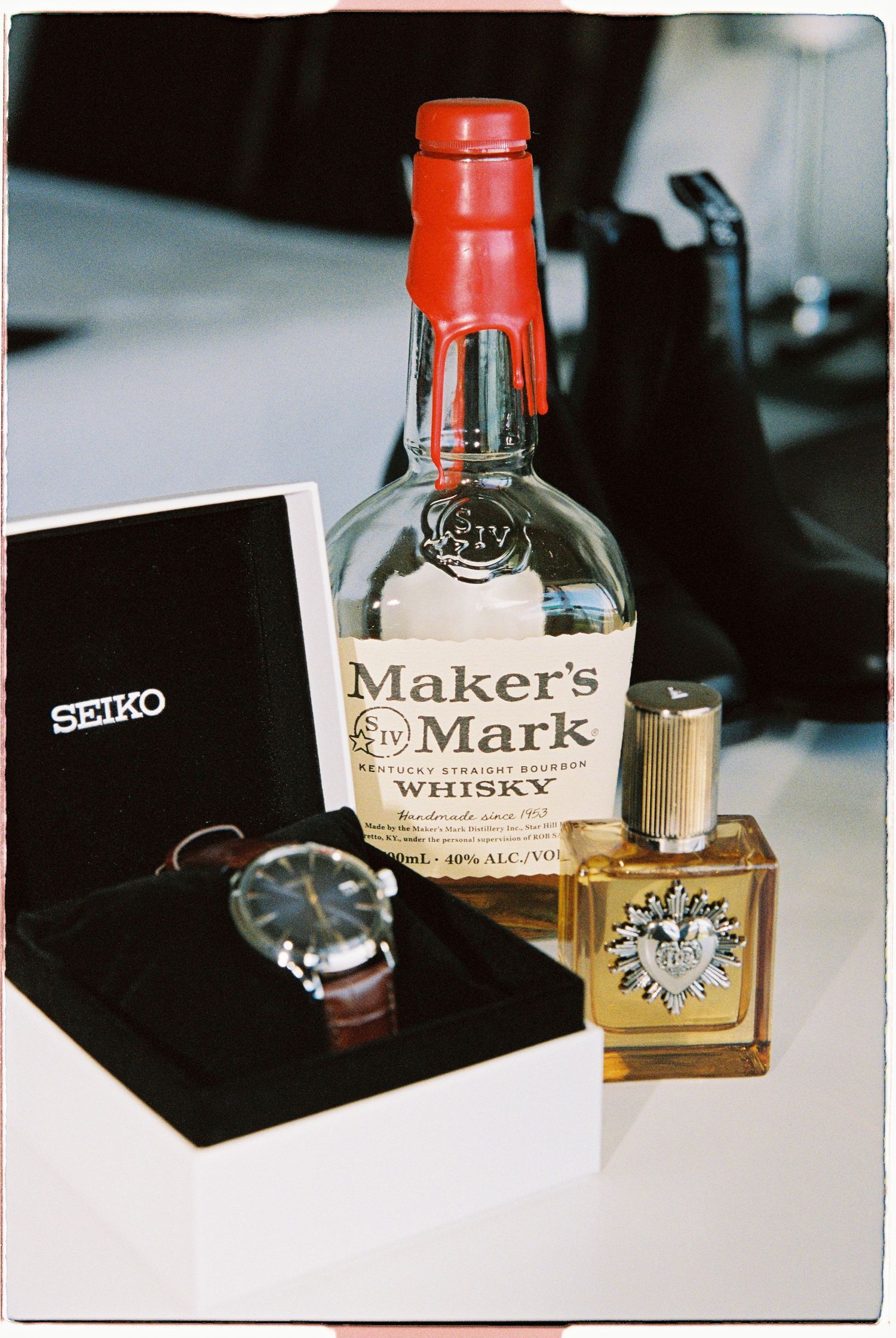 Seiko watch in box beside Makers Mark whisky bottle and gold cologne on a white table.