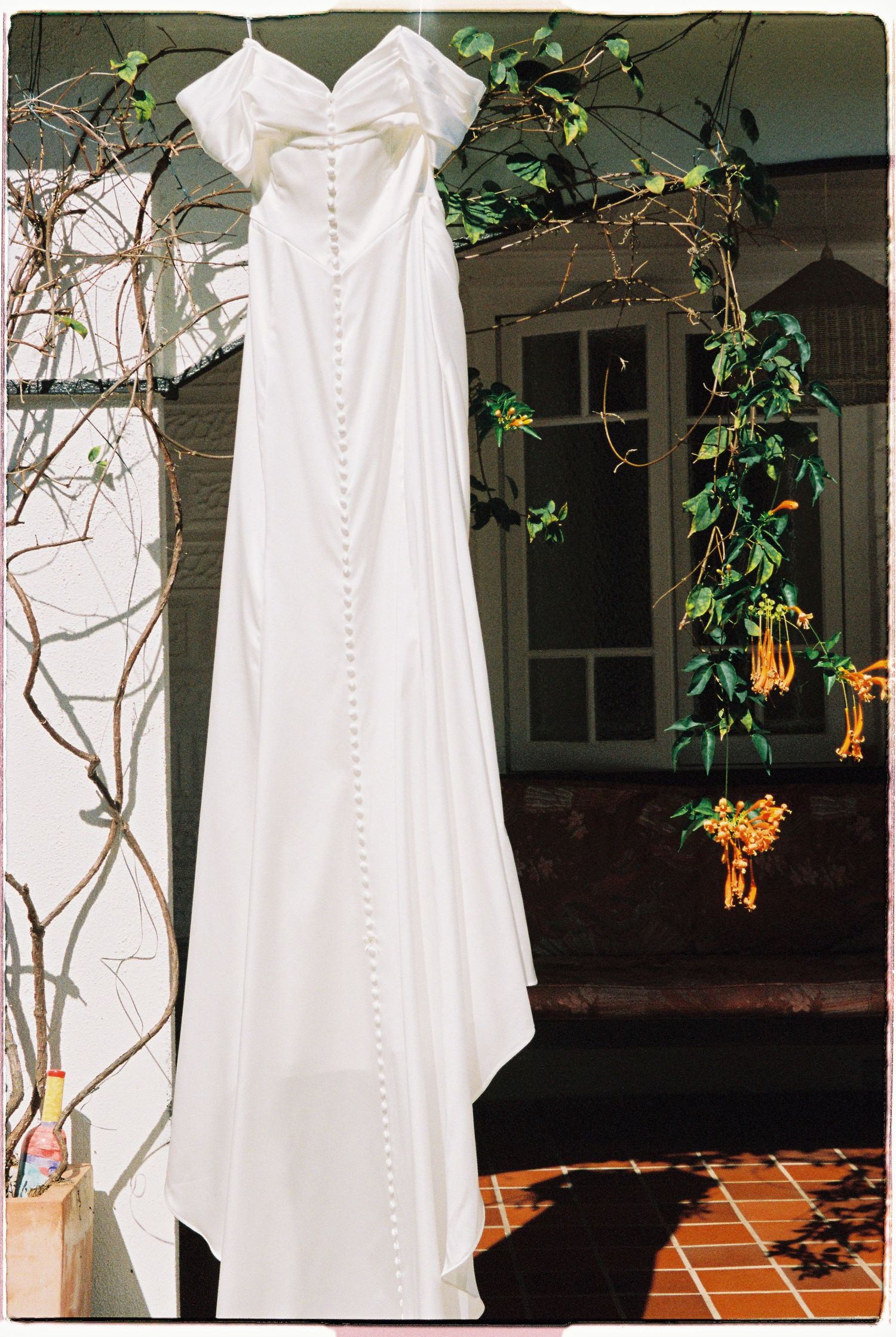Elegant white off-shoulder wedding gown with button back detail hanging outdoors against lush green foliage.