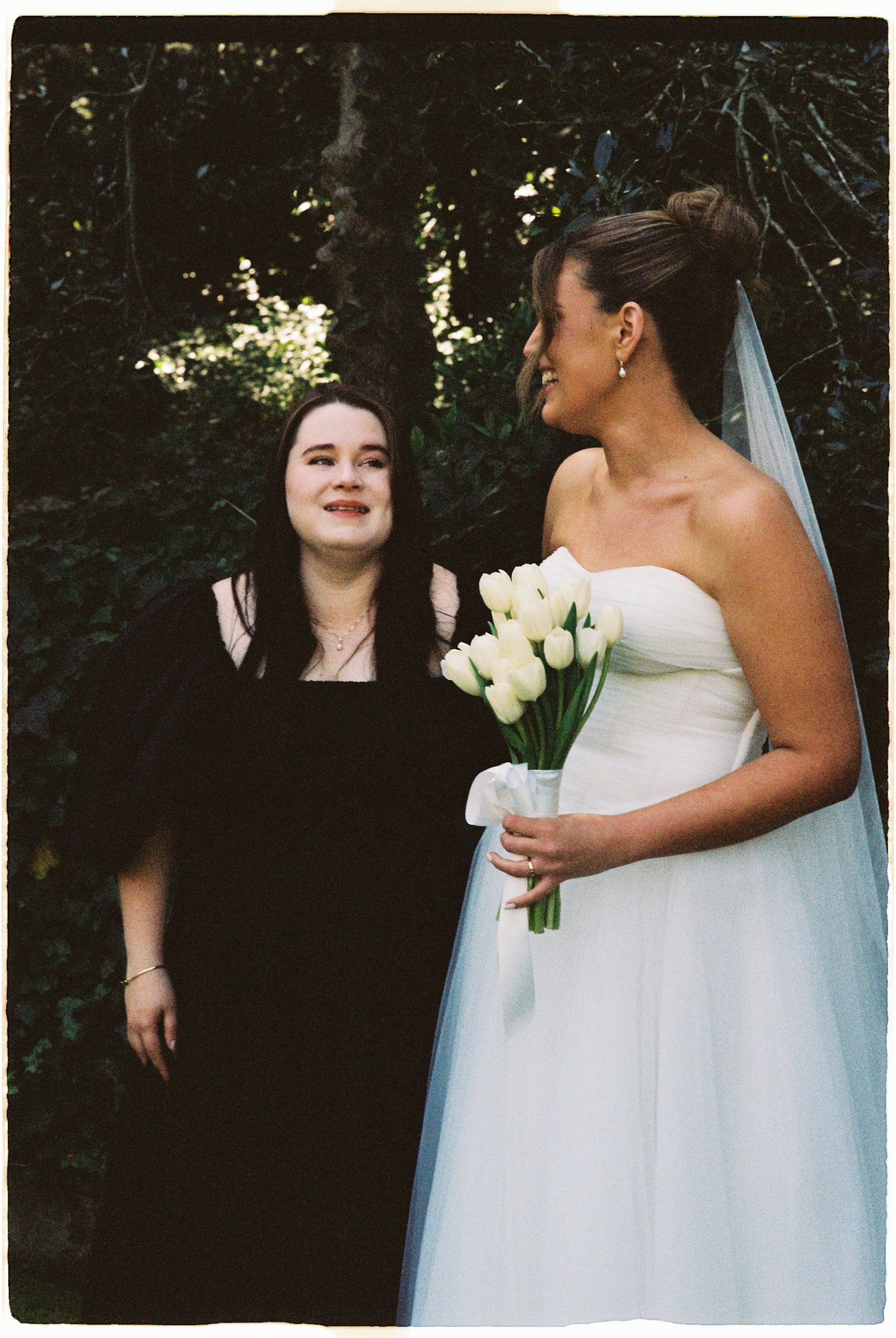 Bride in white strapless gown holding white tulip bouquet stands with guest in black dress outdoors.