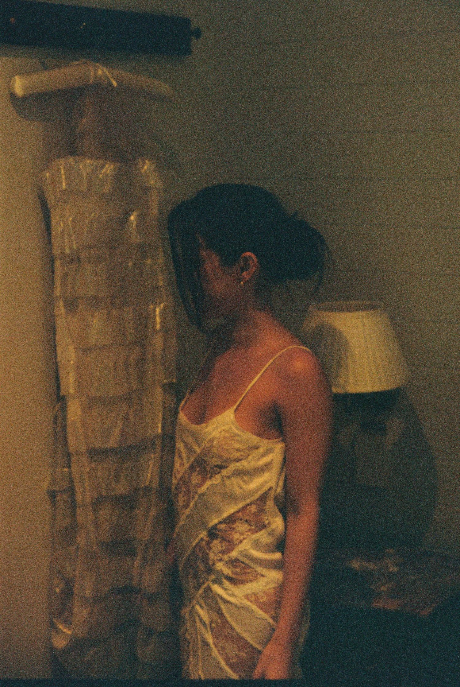Bride in a floral slip dress standing beside her bagged wedding gown in a warmly lit room.