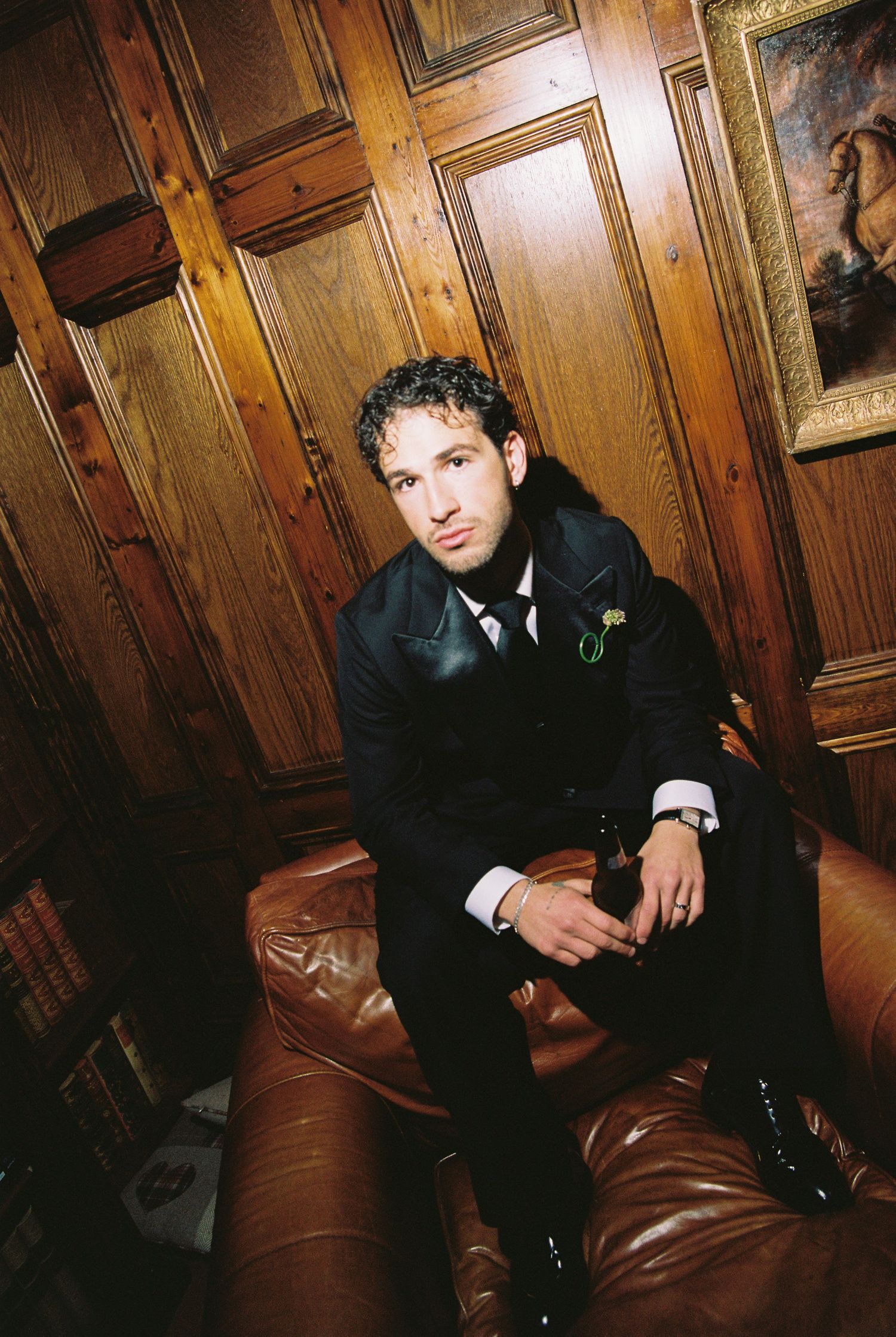 Groom in black suit sitting on leather chair in wood-paneled room with boutonniere.