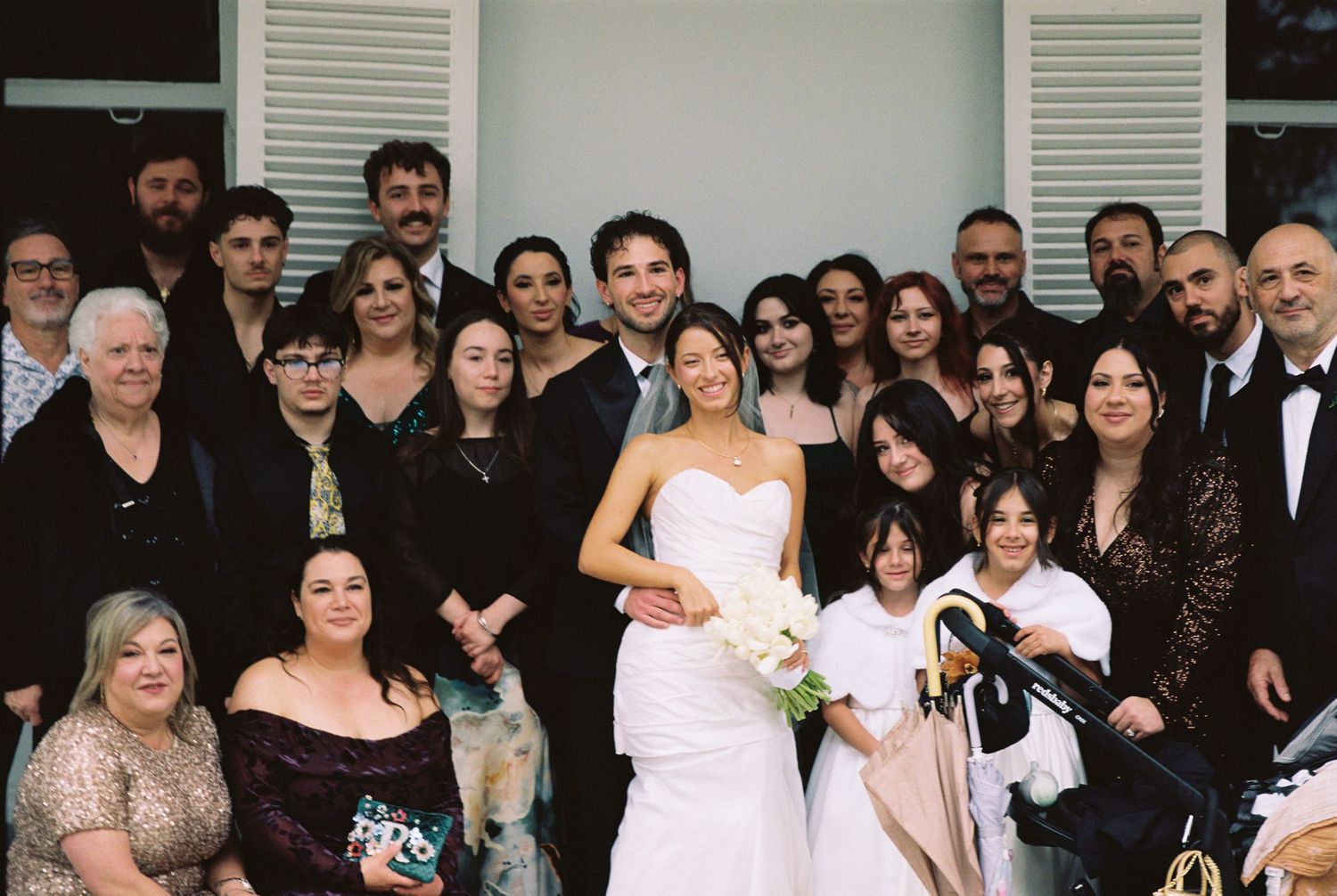 Bride and groom pose with large family group outside elegant white wedding venue.