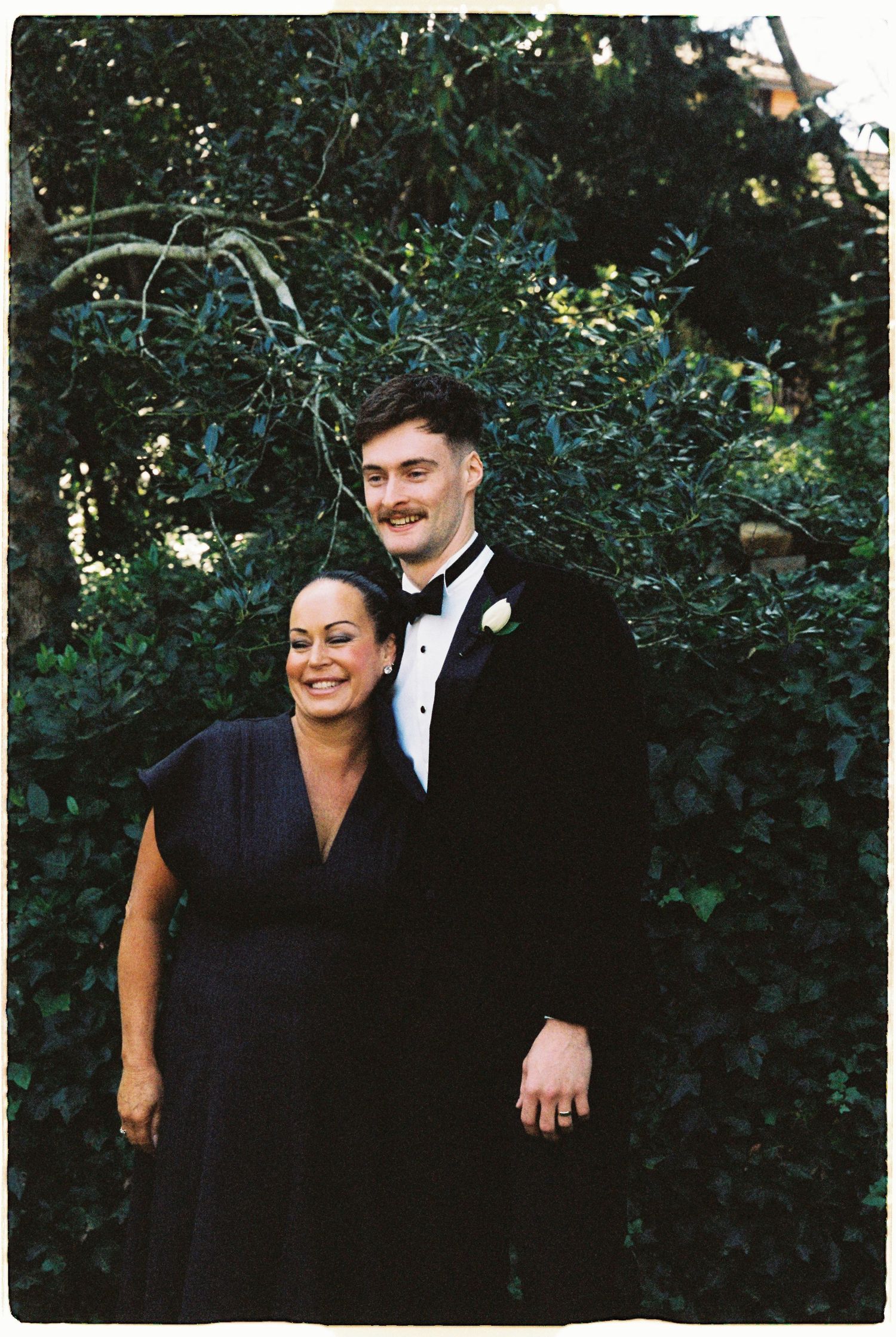 Groom in black tuxedo posing with woman in navy dress against green foliage.