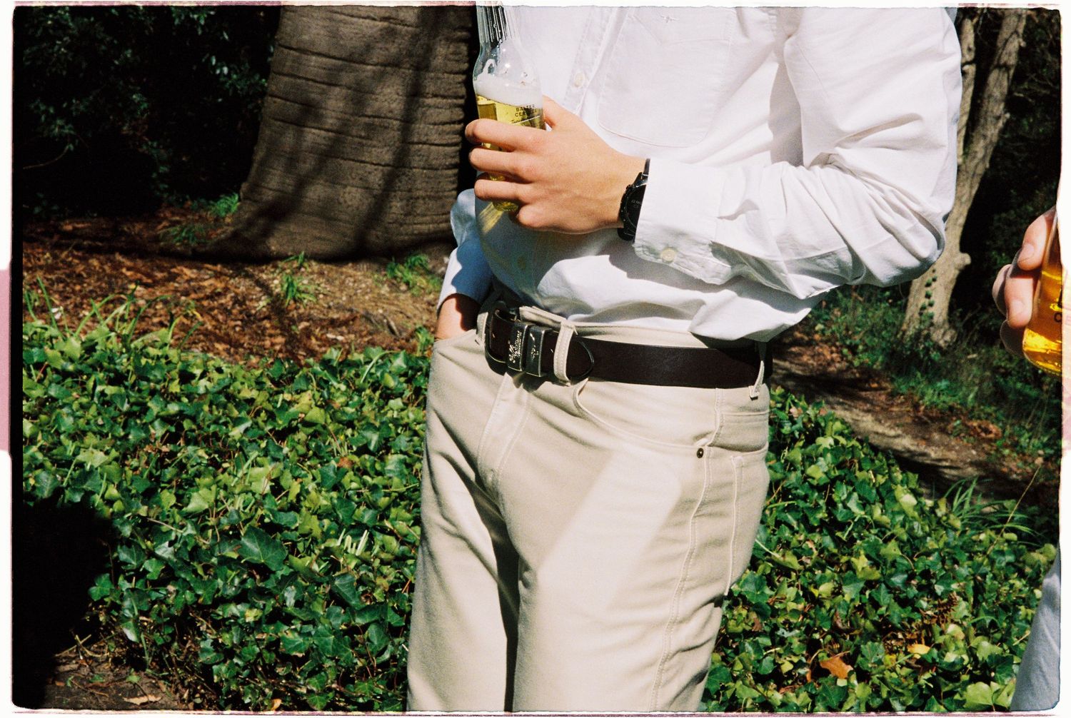 Man in white dress shirt and khaki pants holds a drink while standing in a garden at a wedding.