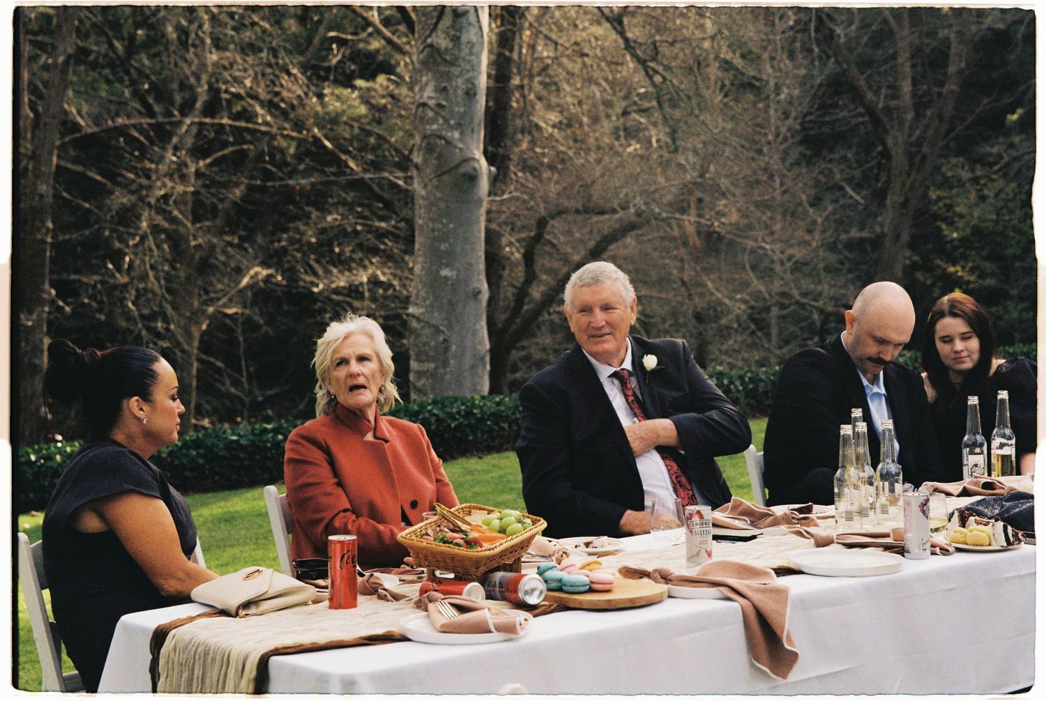 Wedding guests seated at outdoor reception table surrounded by lush green trees in garden setting.