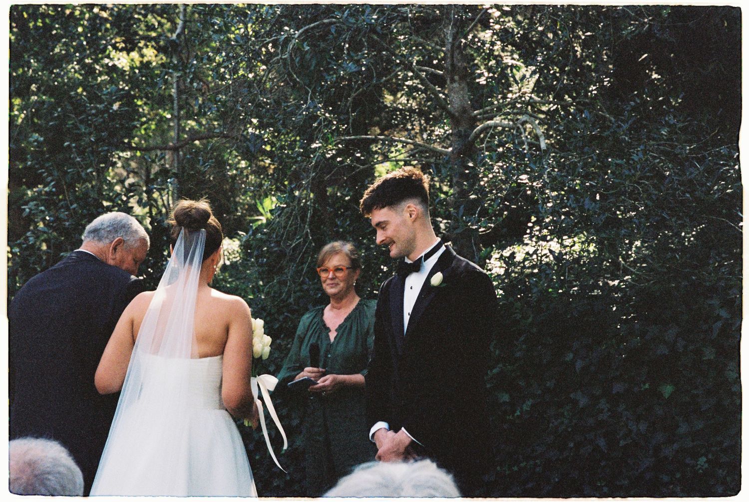 Groom in black tuxedo standing at outdoor wedding ceremony altar with bride and officiant.