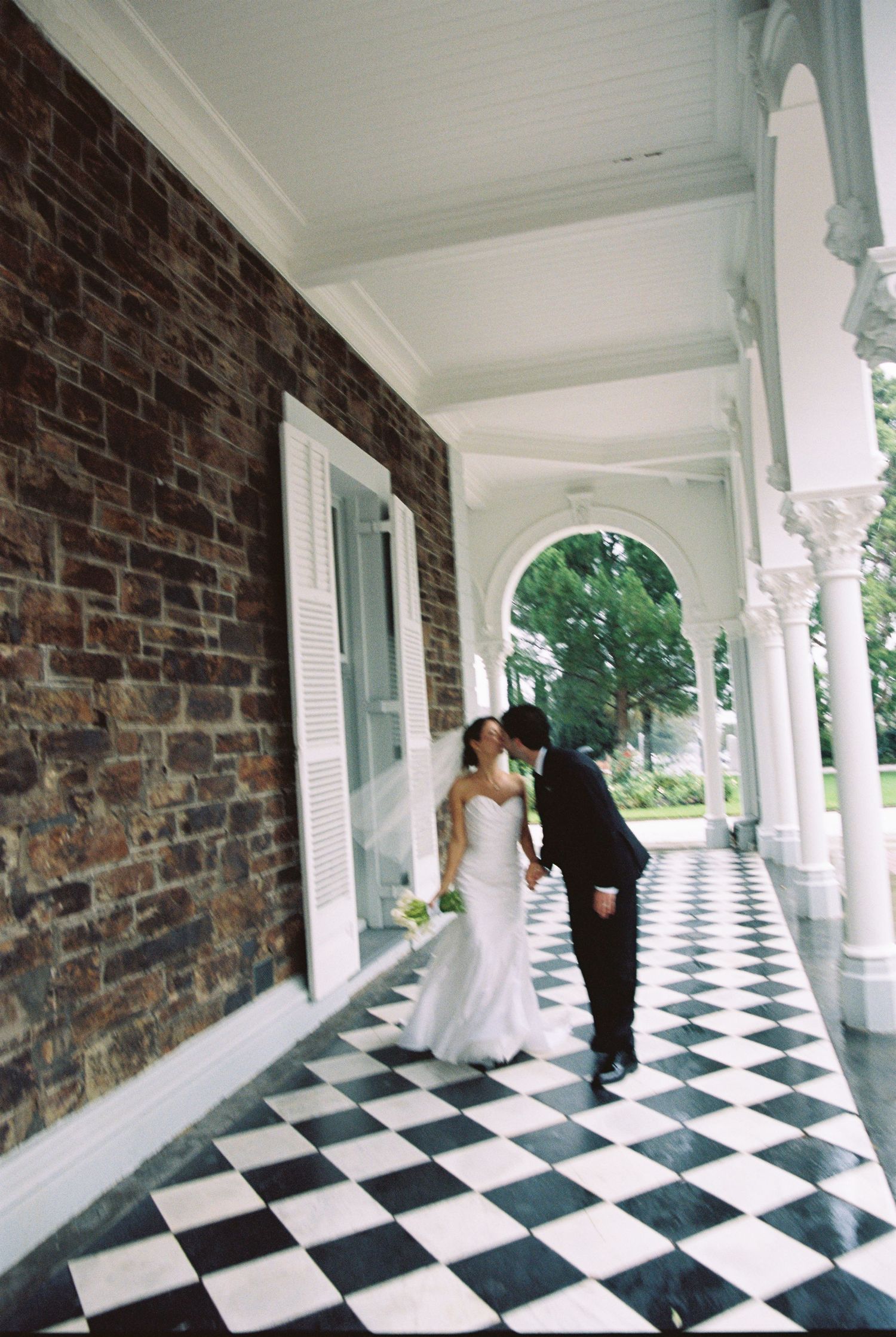 Bride and groom share a kiss on a classic black and white checkered porch of a historic venue.