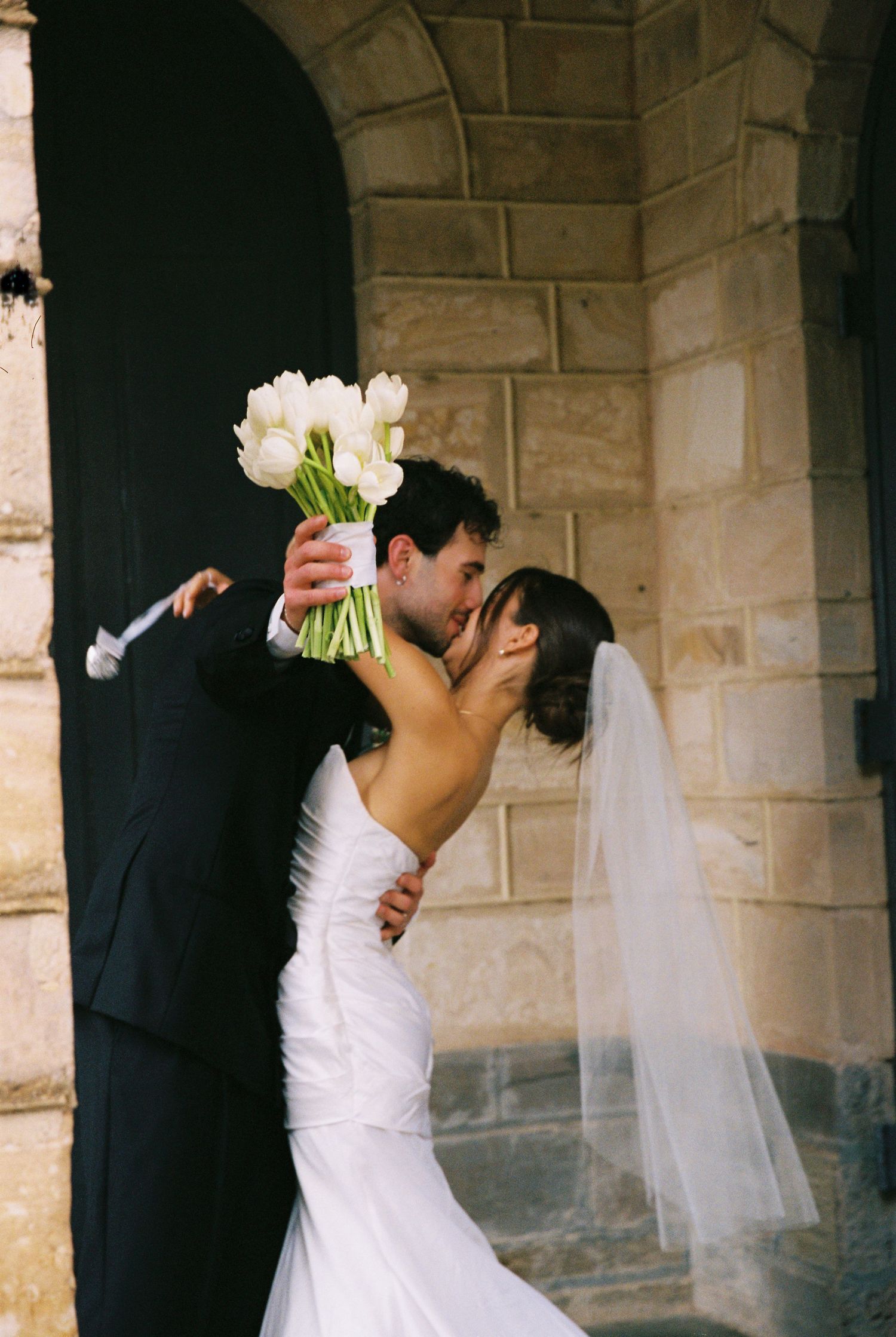 Groom dips bride for romantic kiss outside stone church doorway, holding white tulip bouquet.