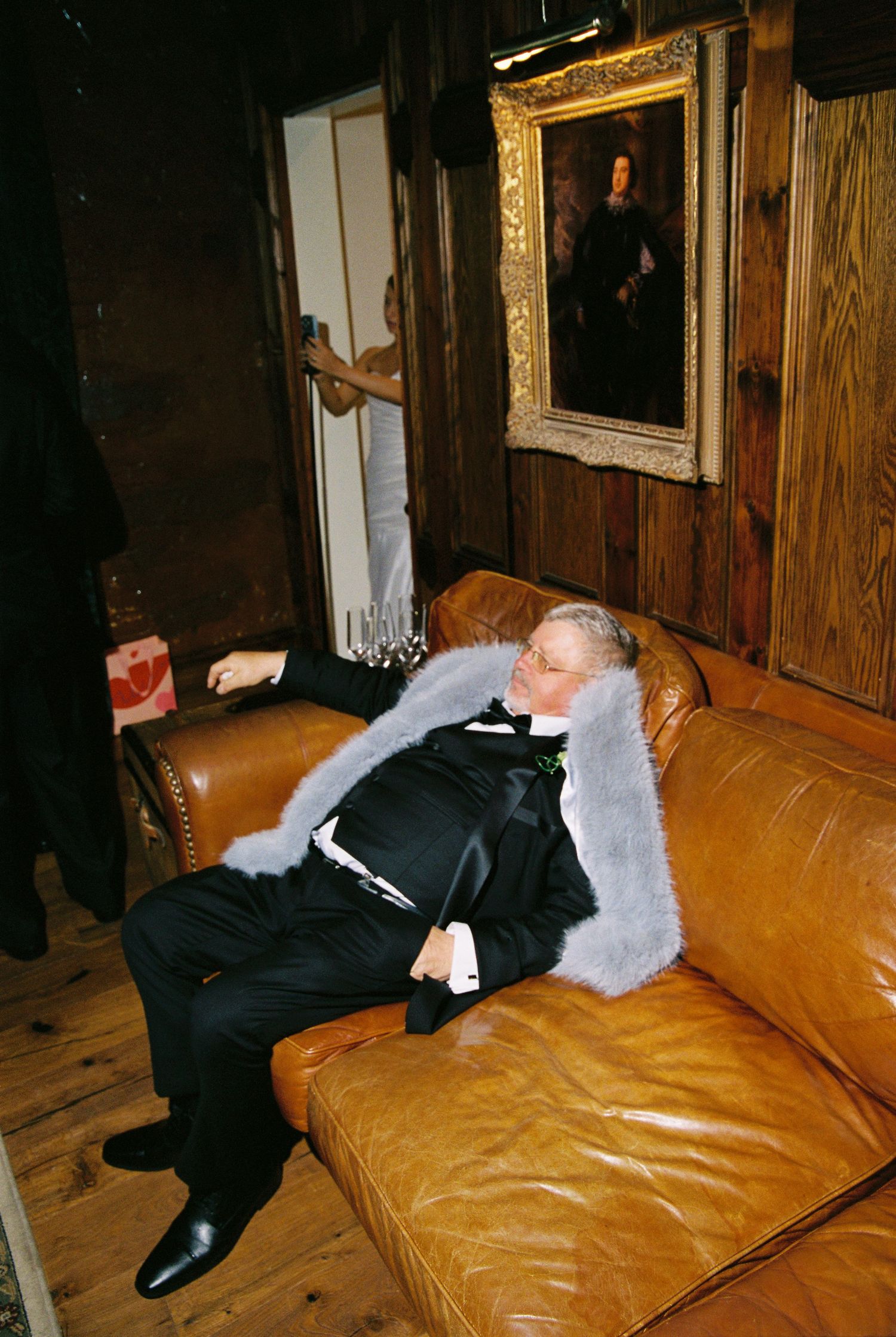 Man in tuxedo with fur stole relaxing on leather sofa beneath a classic portrait painting.