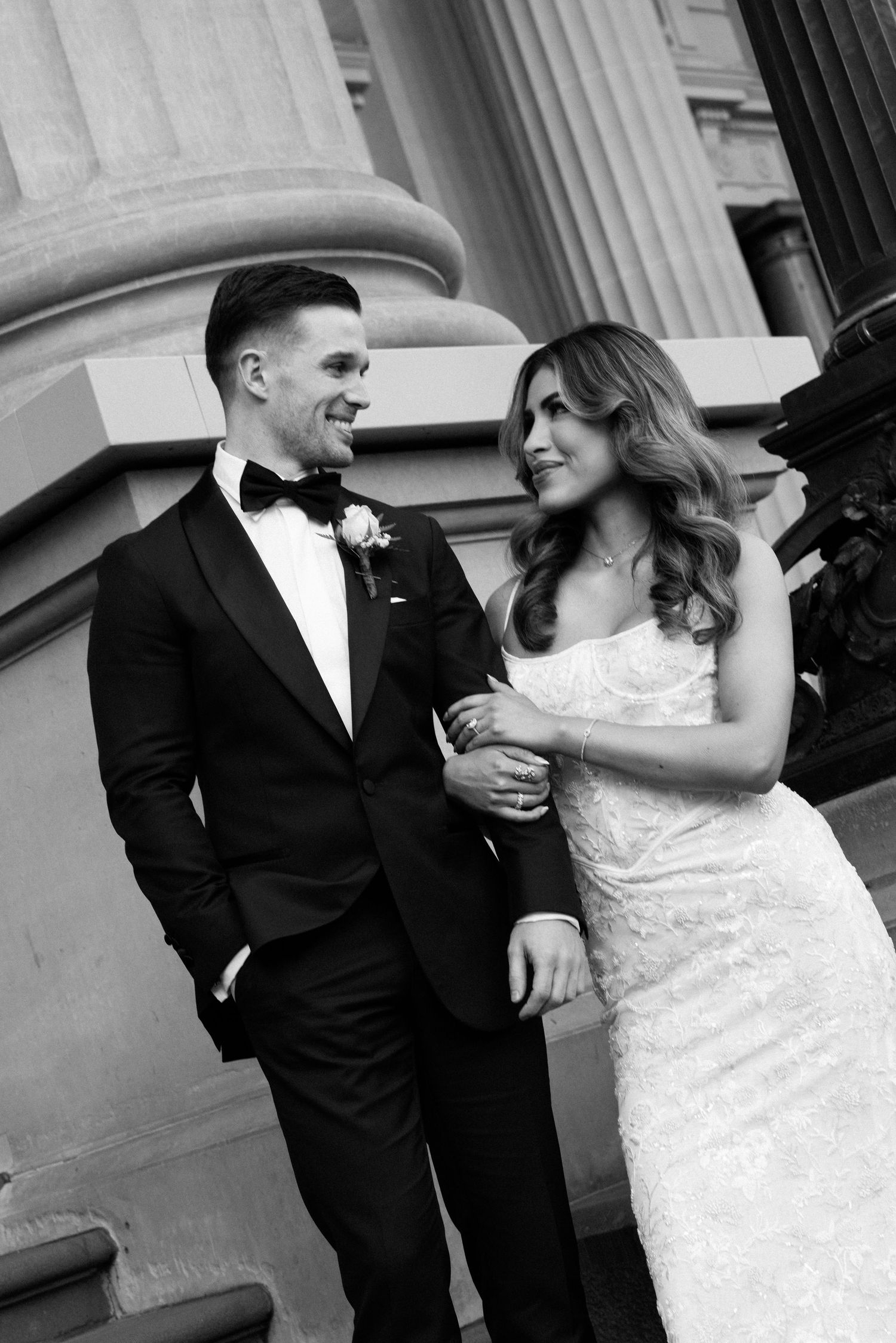 Black and white photo of a bride and groom smiling at each other beside grand stone columns.