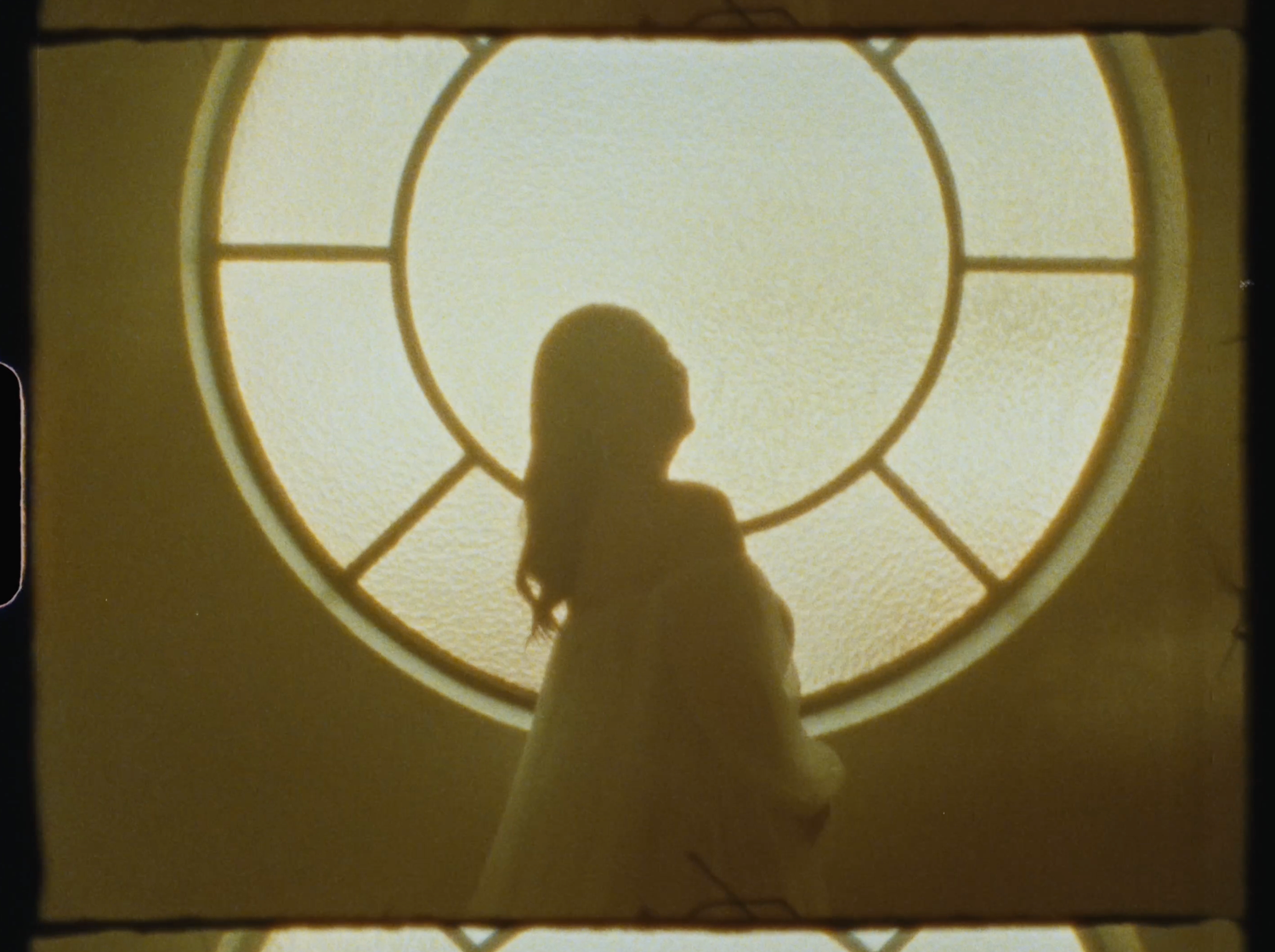 Silhouette of a woman standing before a large circular window, bathed in warm golden light.