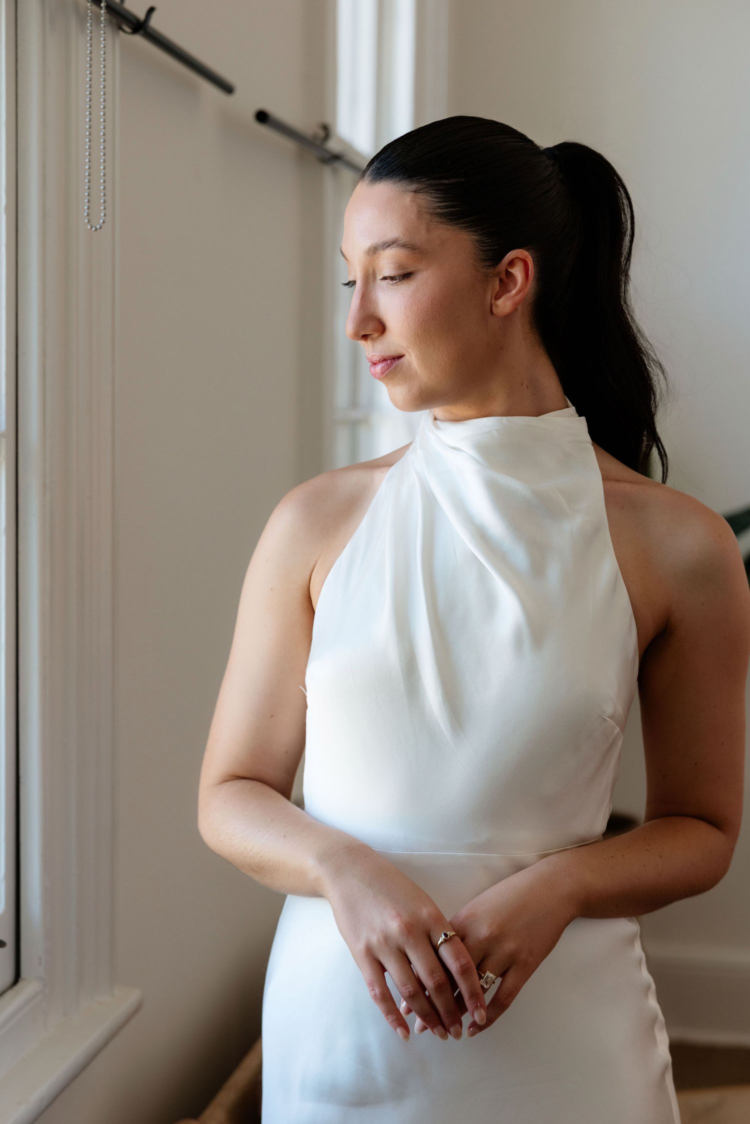 Bride in elegant white halter neck wedding dress standing by window with eyes closed, wearing ring.