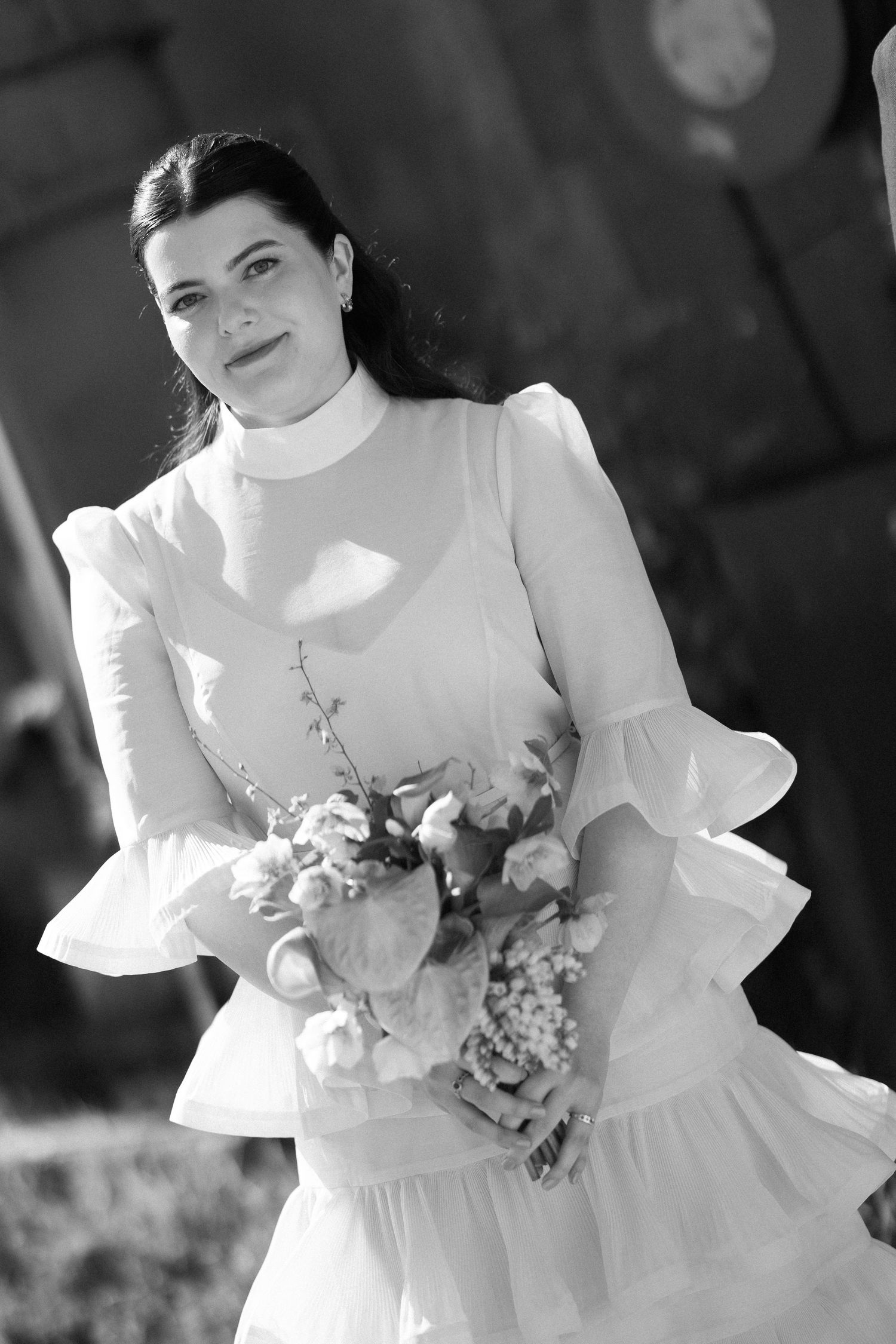 Black and white portrait of dark-haired bride in ruffled dress holding floral bouquet outdoors.