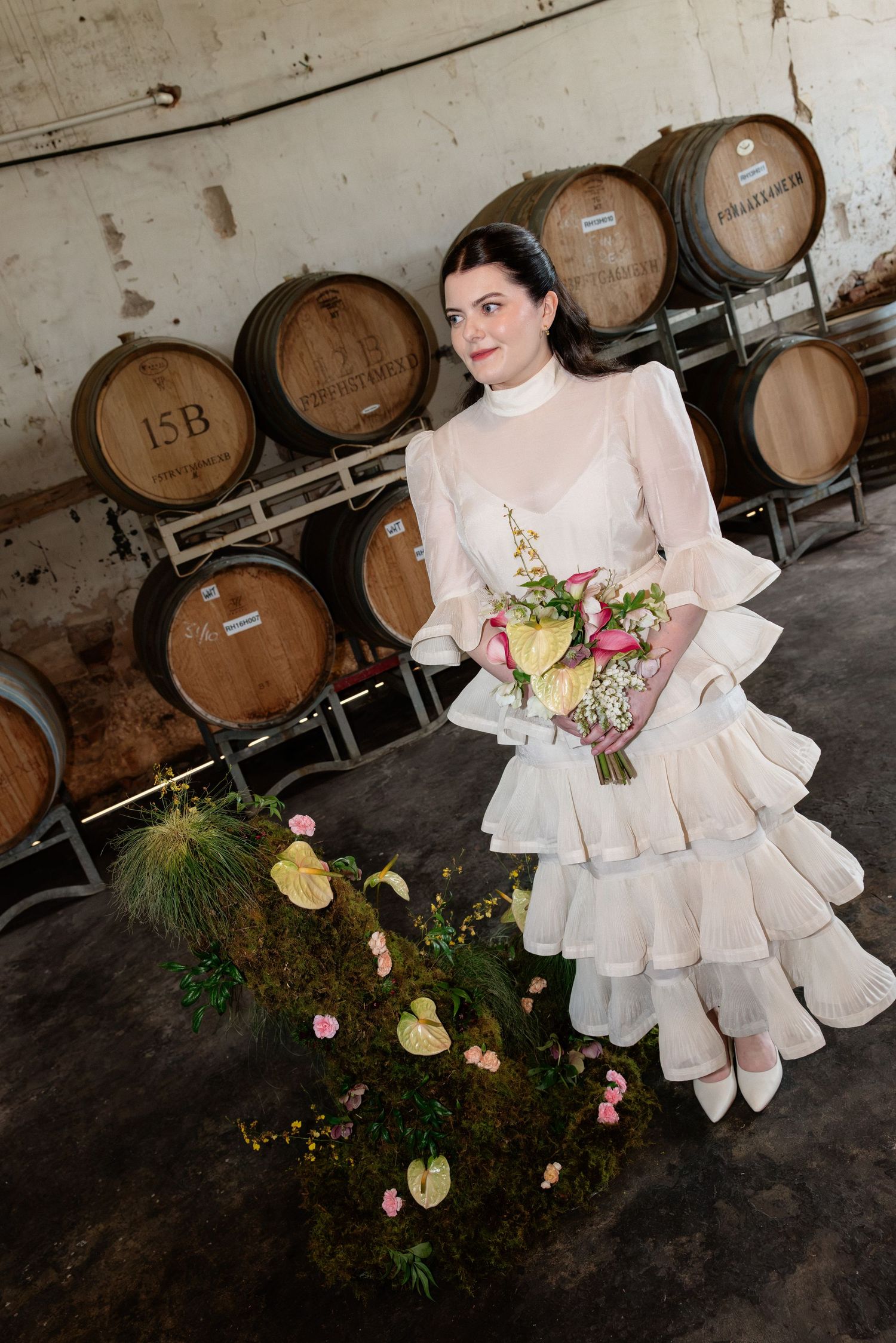 Bride in tiered white ruffled dress holds tropical bouquet beside wine barrels in a rustic winery setting.