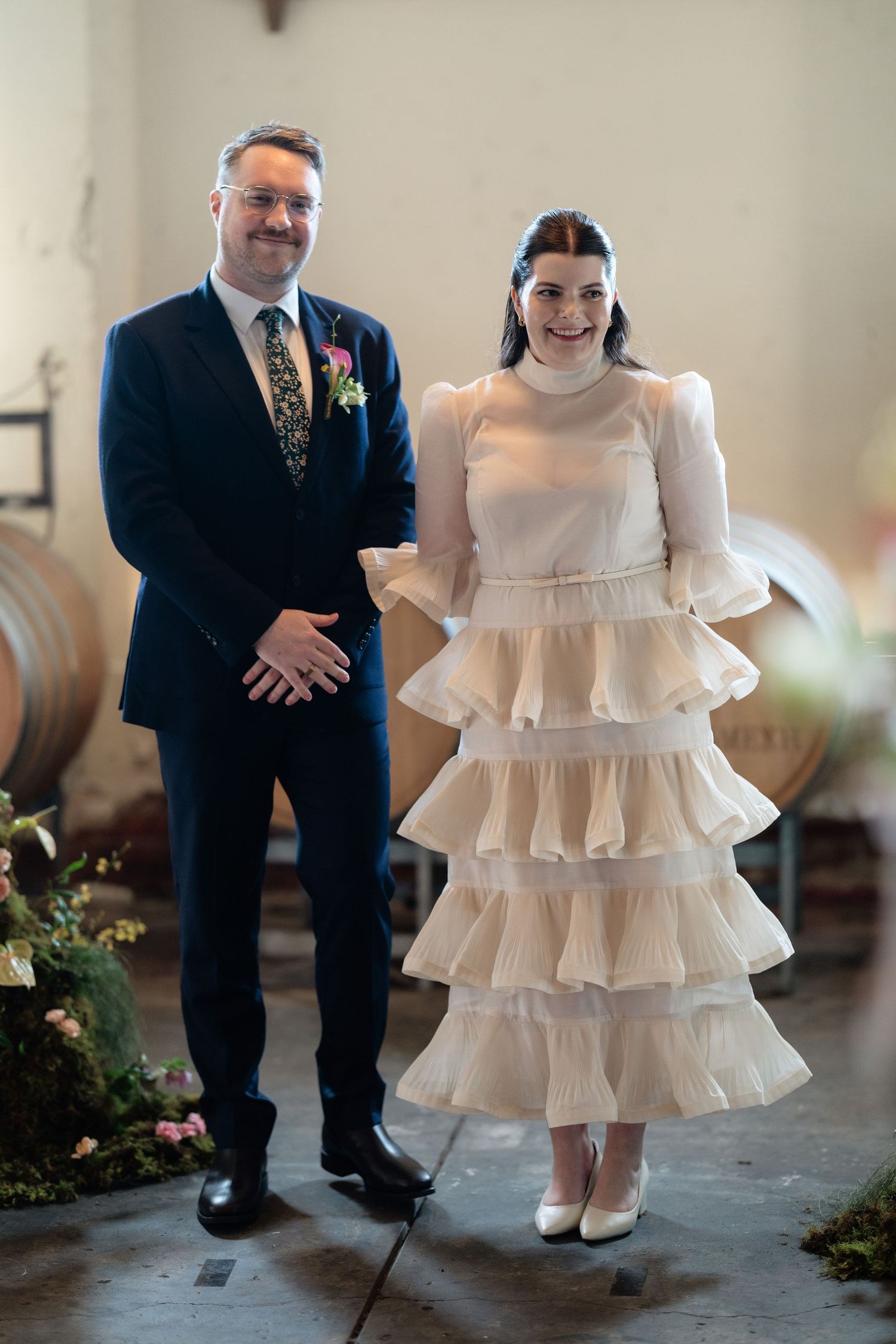 Couple posing in winery with oak barrels, bride in tiered cream ruffle dress holding bouquet, groom in navy suit.