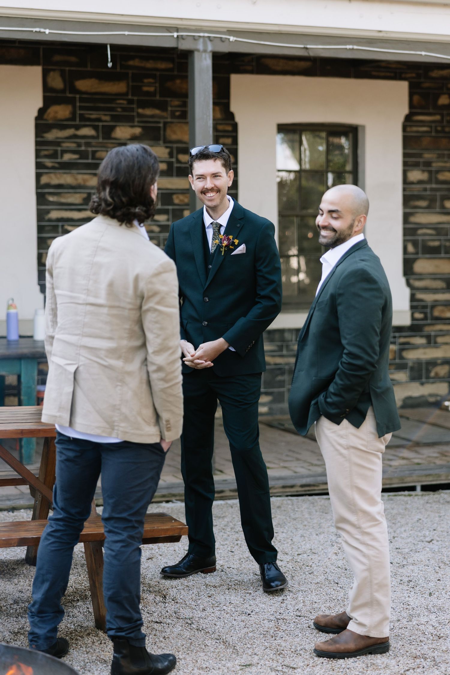 Three men in semi-formal attire laugh and converse outdoors at a wedding reception near a stone building.