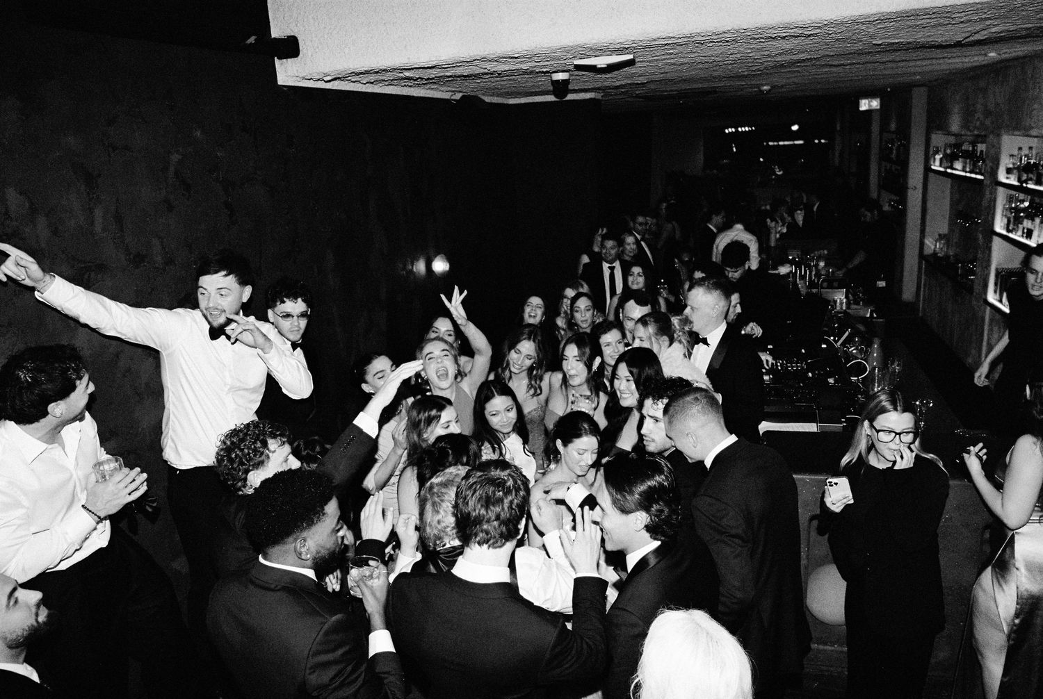 Lively wedding reception with guests dancing and celebrating in dimly lit venue.