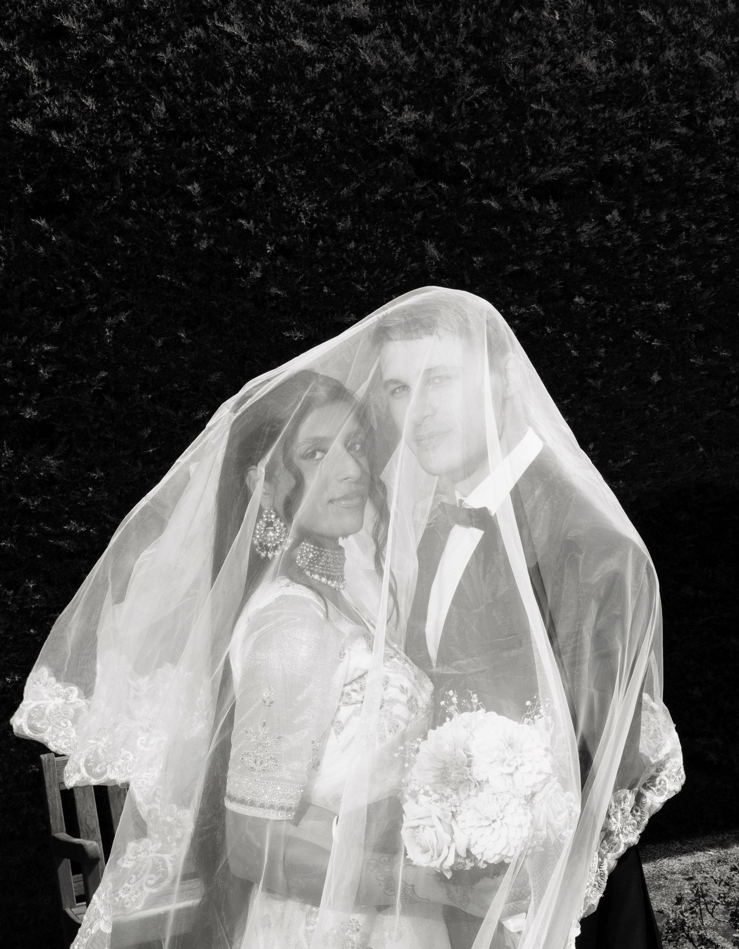 Dramatic black and white wedding portrait of bride and groom posed beneath a flowing lace veil outdoors.