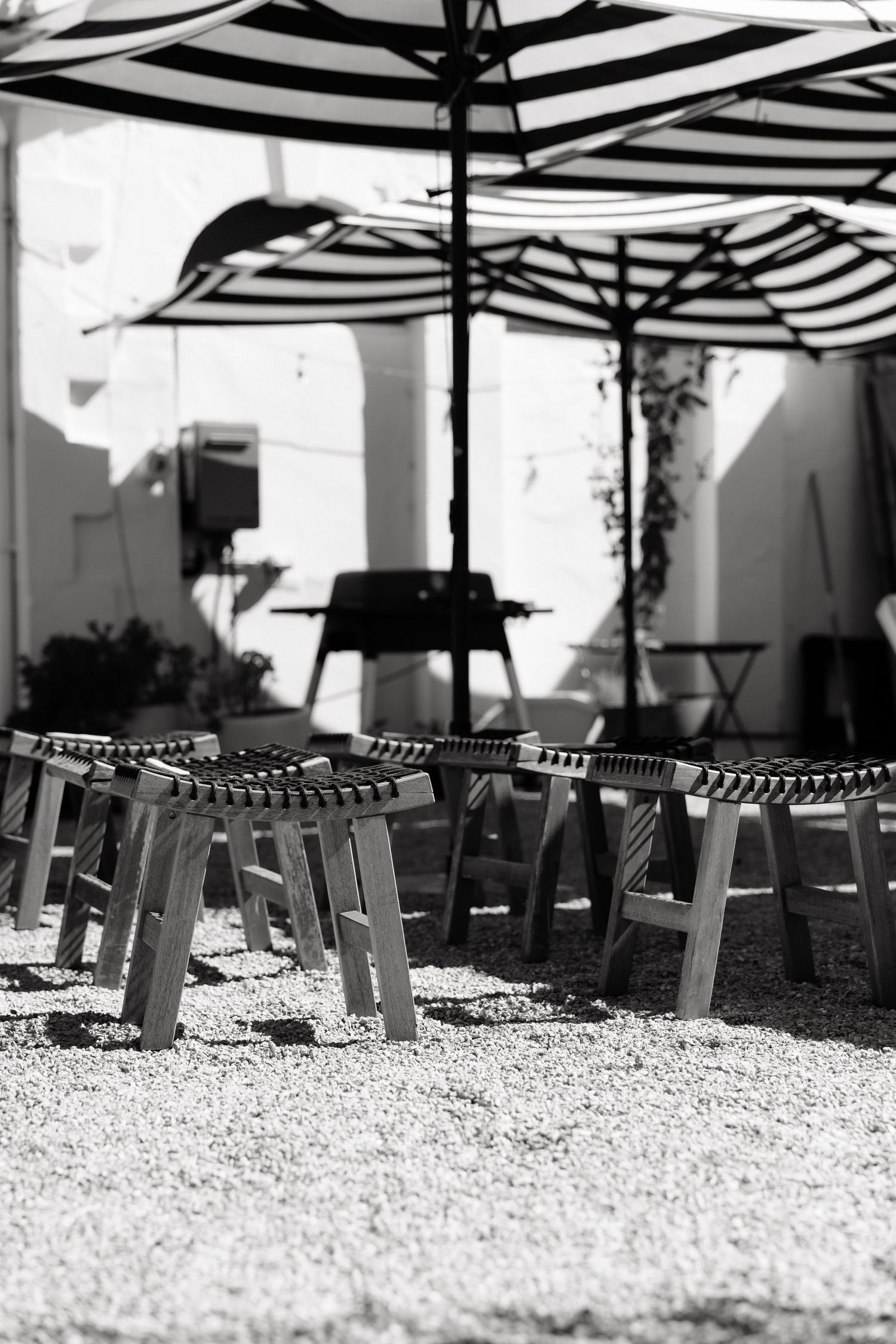 Wooden stools arranged under striped black and white outdoor umbrellas on a gravel patio.