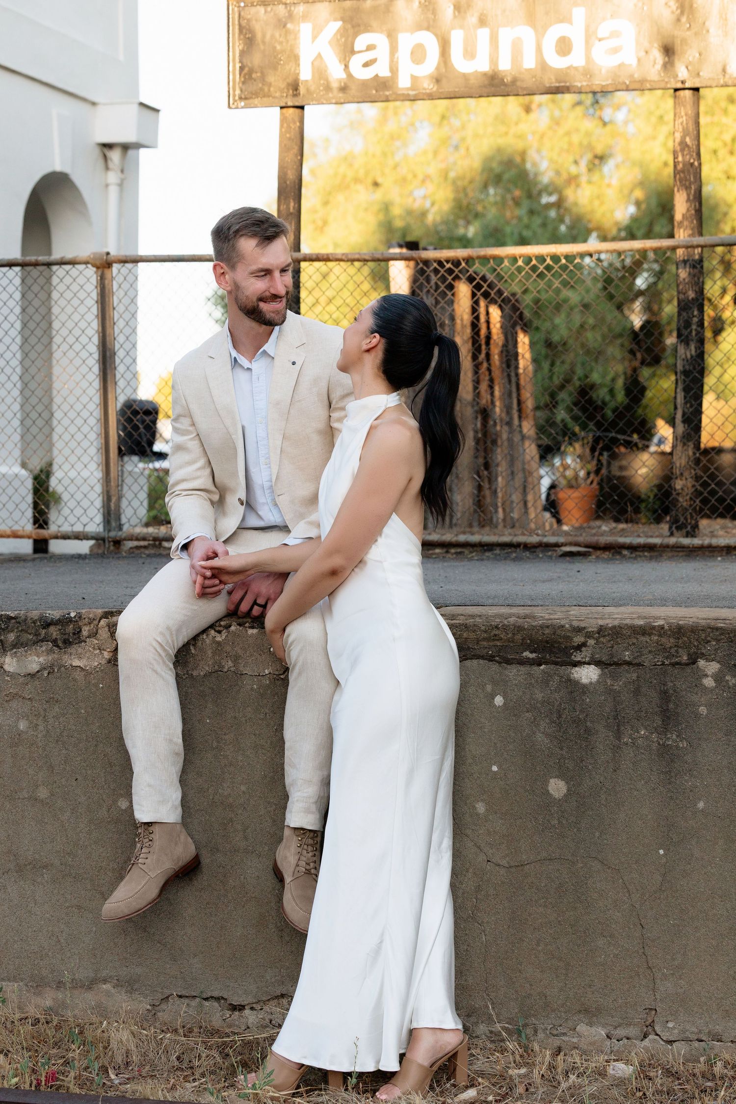 Bride and groom pose together near vintage Kapunda railway sign, groom seated on stone wall in beige suit.