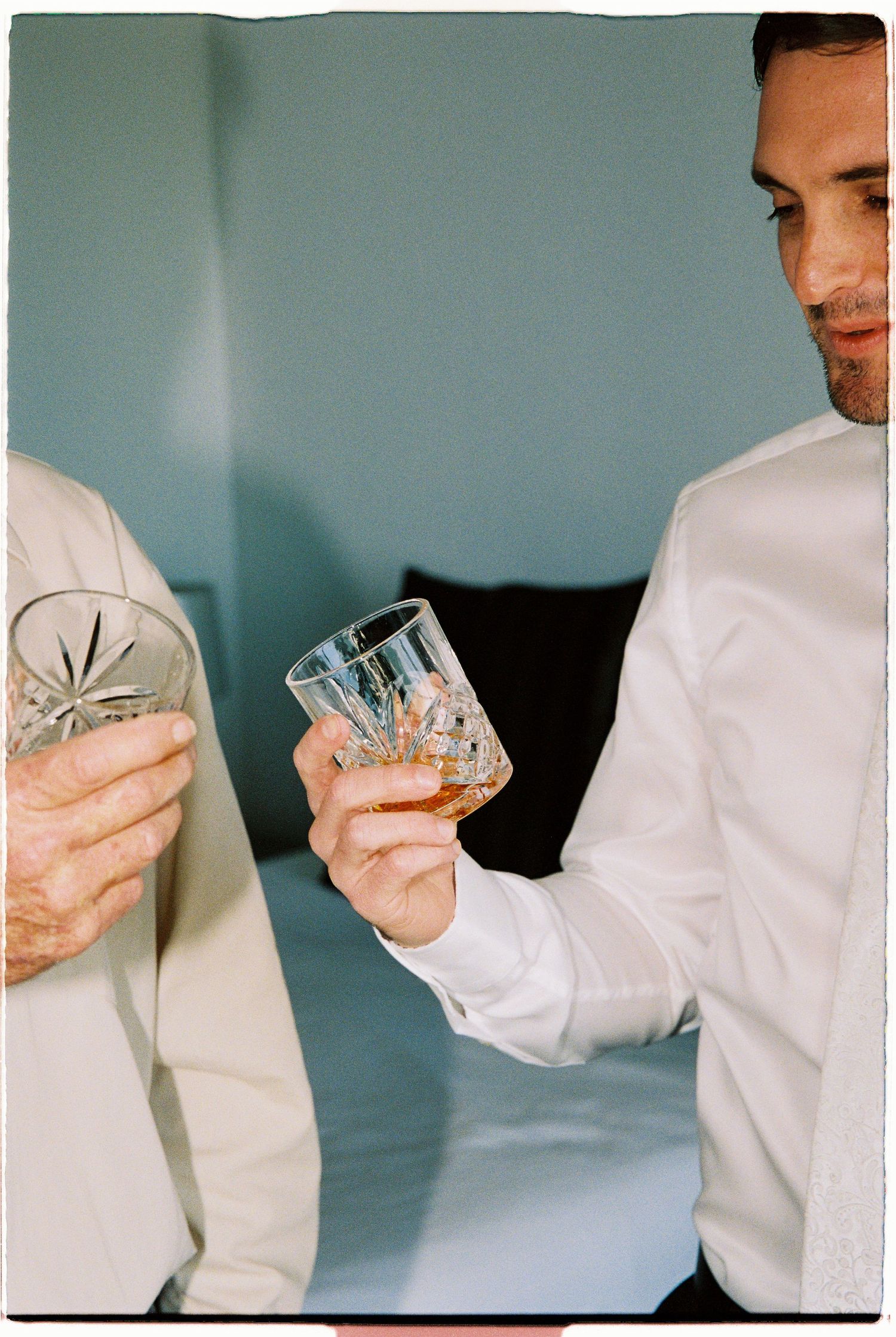 Two men in white shirts share a toast with crystal whiskey glasses at a wedding celebration.