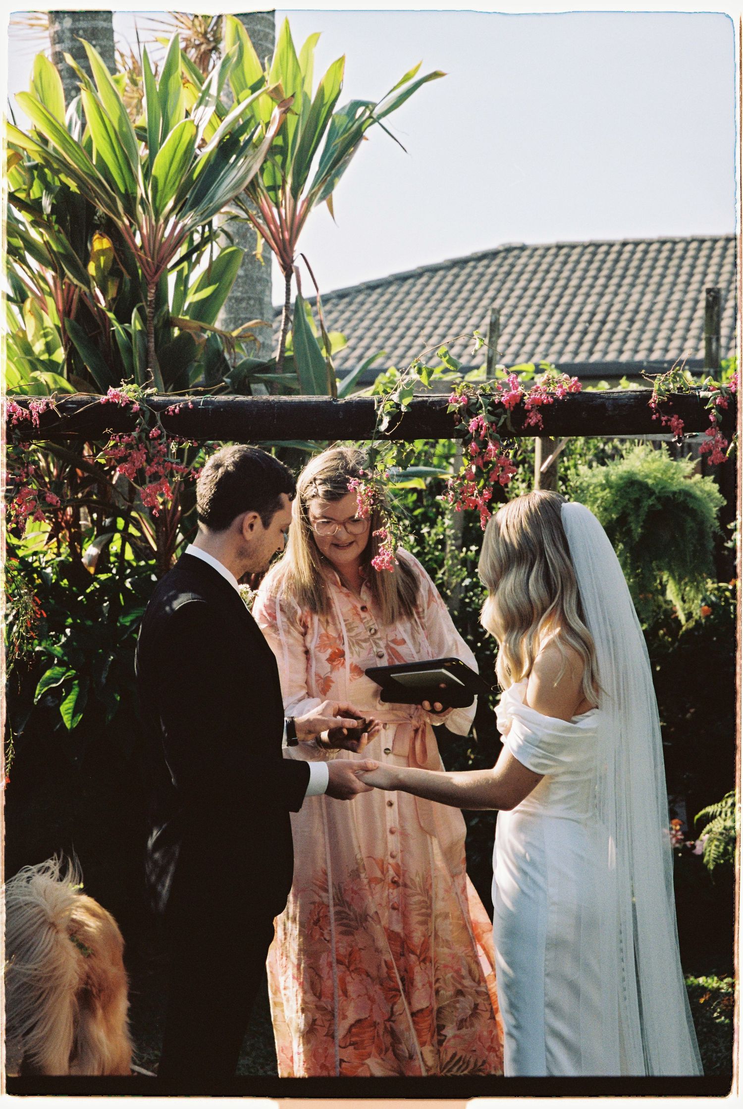 Officiant in floral dress leads outdoor wedding ceremony with bride in white gown and groom exchanging rings.