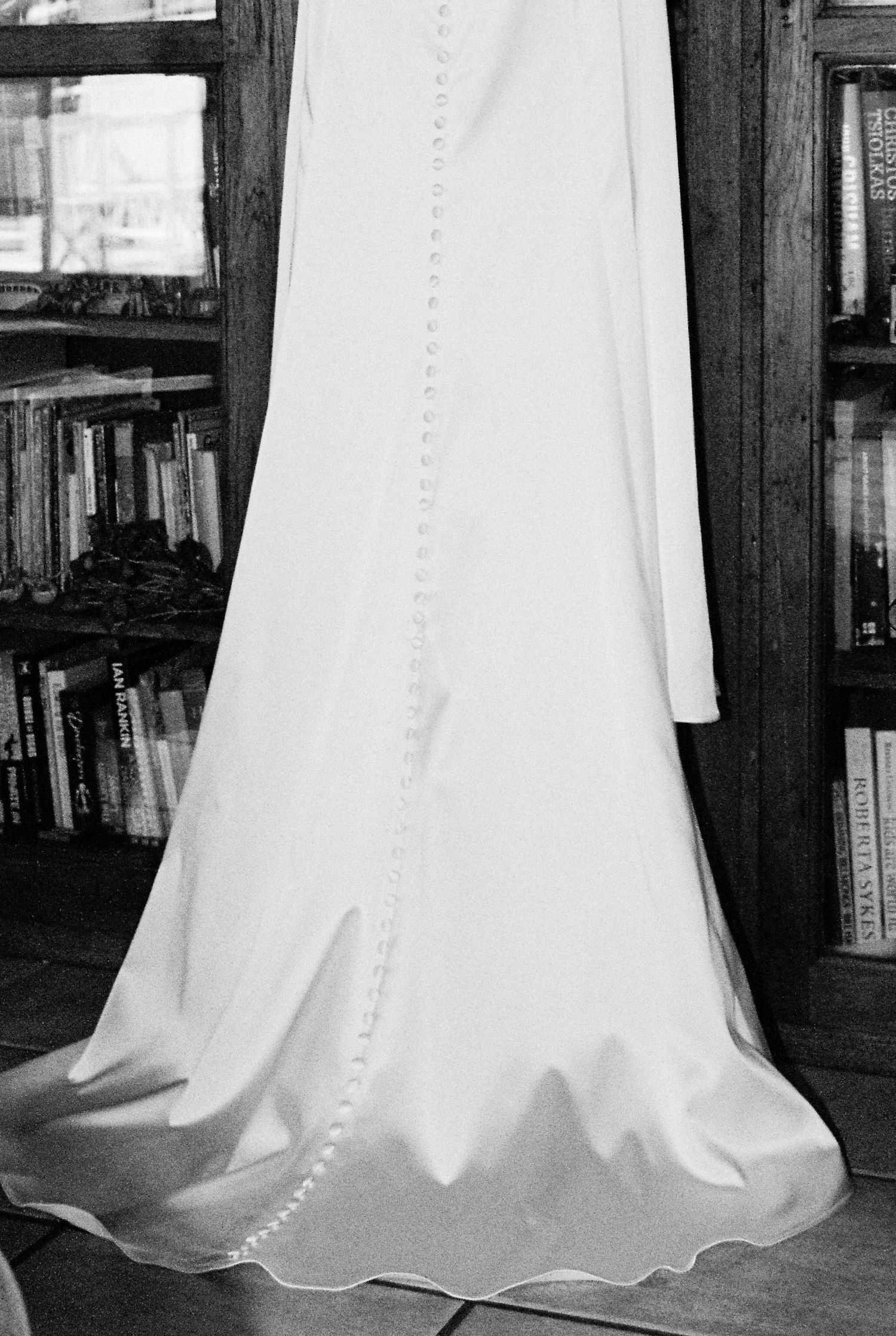 Black and white close-up of white wedding dress with button back detail hanging near bookshelf.
