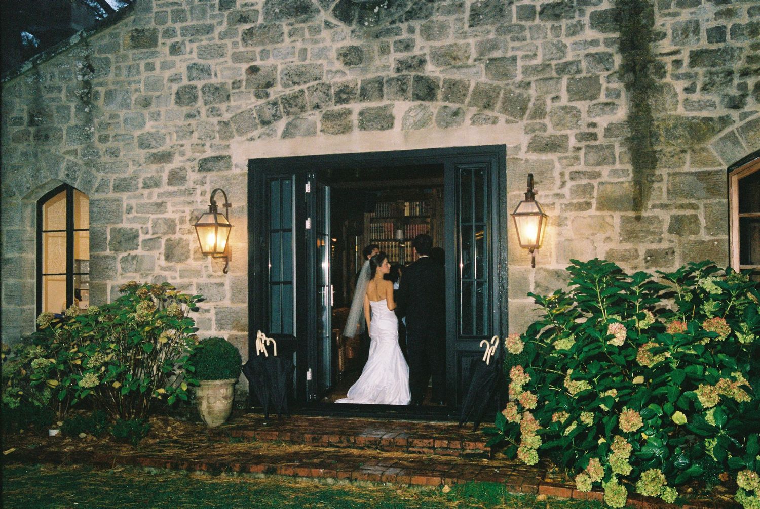 Bride in white wedding gown standing in doorway of rustic stone venue with lanterns at dusk.