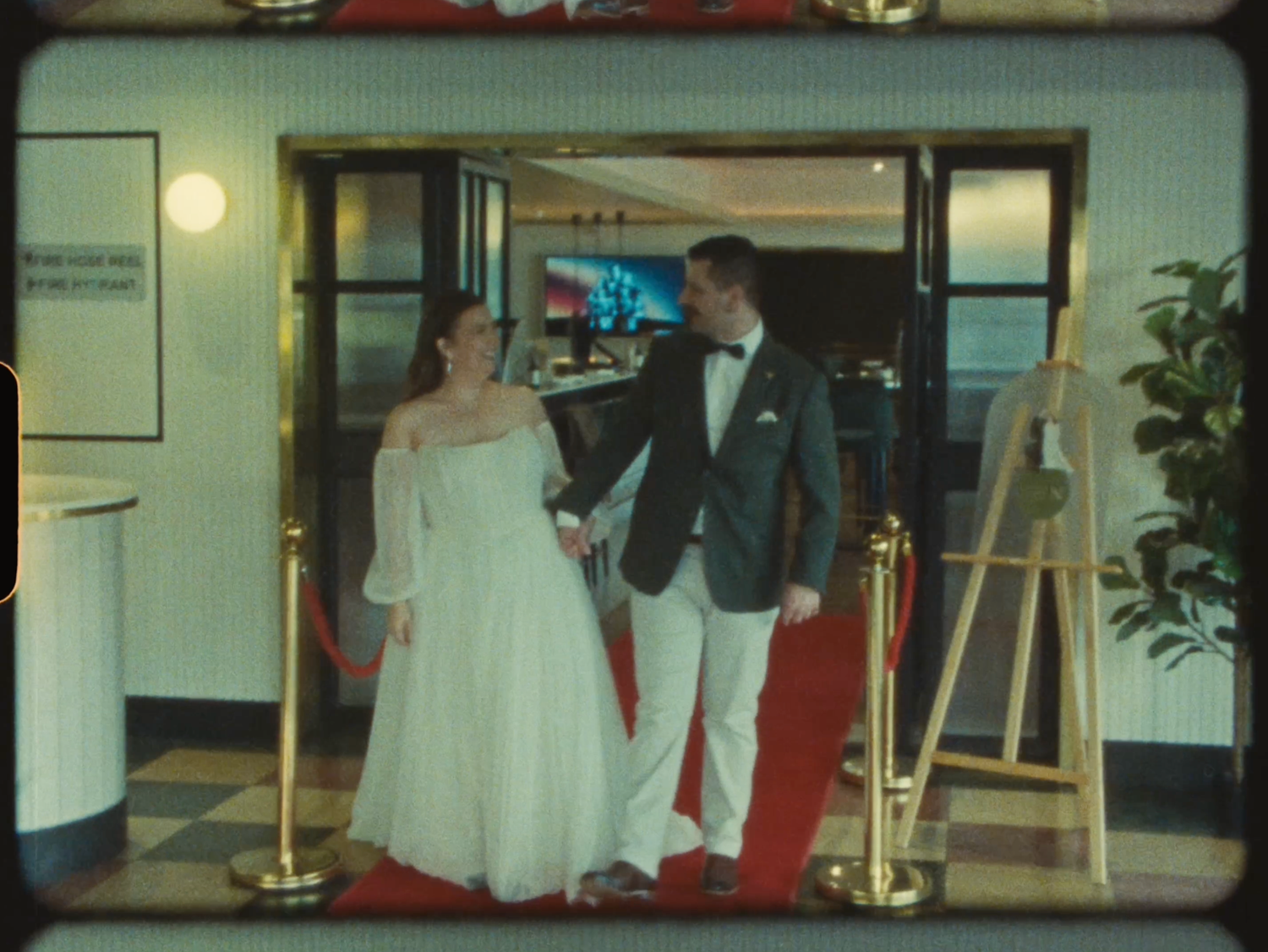 Bride and groom walking hand in hand on a red carpet through elegant cinema entrance doors.