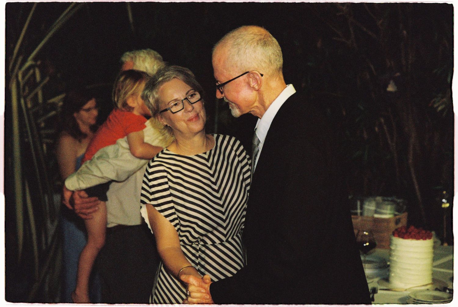 Elderly couple dancing together at an outdoor evening reception, with guests and a white cake visible in the background.