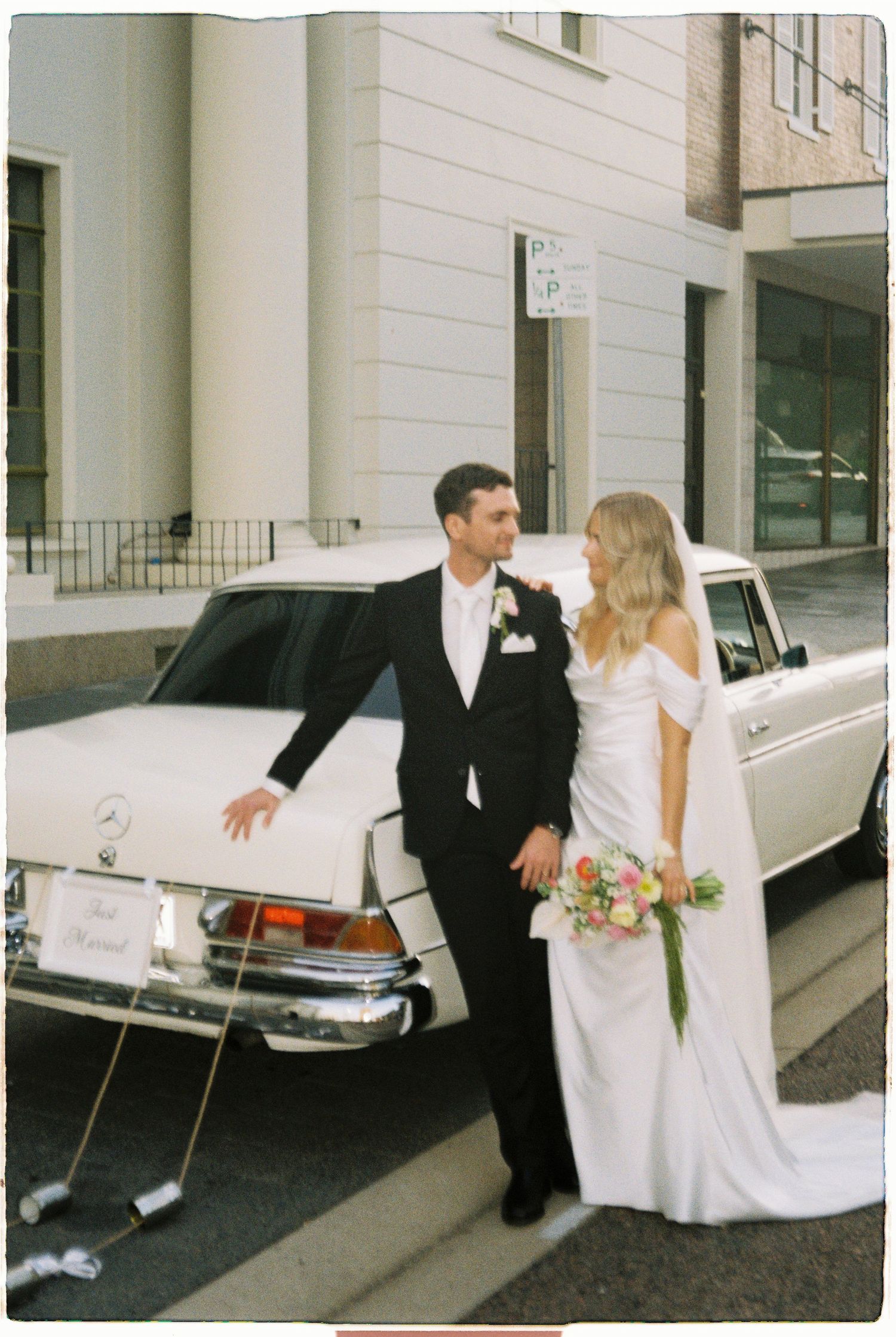 Bride and groom pose beside a vintage white Mercedes wedding car on a city street, bride holding a colorful bouquet.