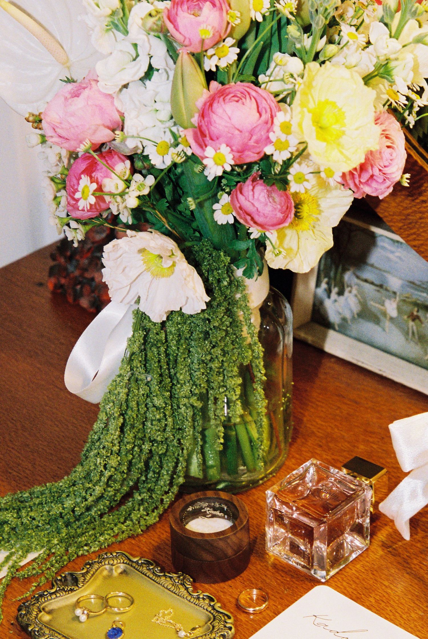 Wedding flat lay with pink and yellow floral arrangement, rings, and accessories on wooden table.