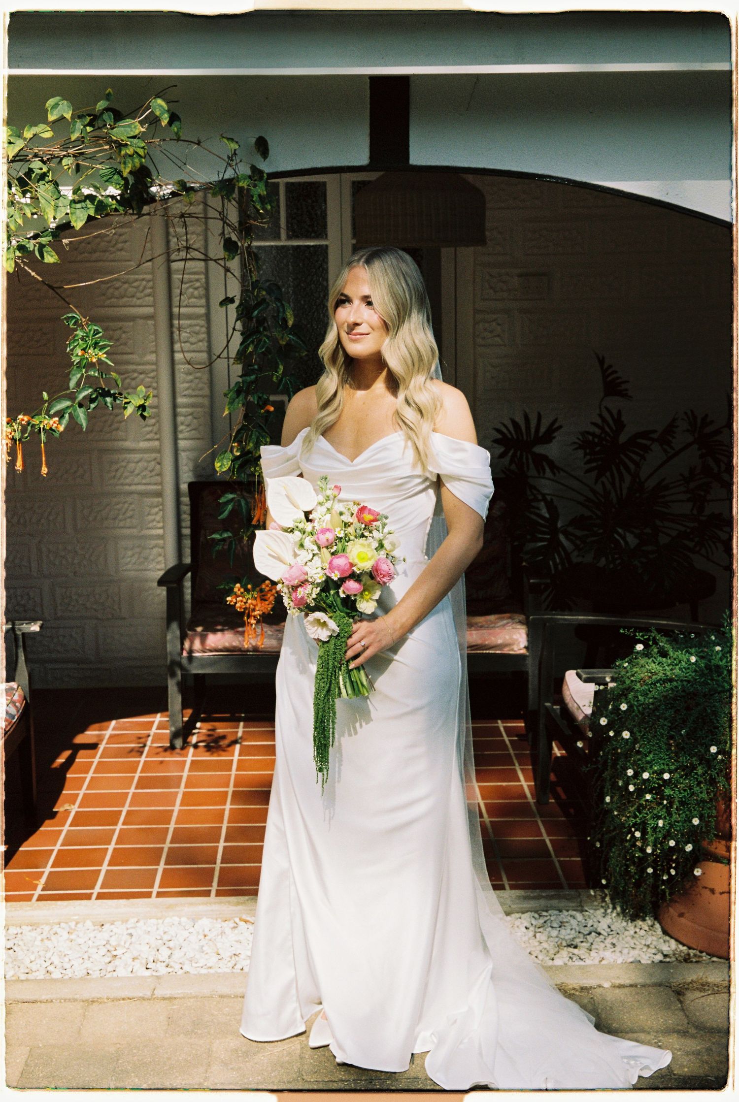 Elegant bride in off-shoulder white gown holding colorful bouquet standing on terracotta tiled porch with arched doorway.