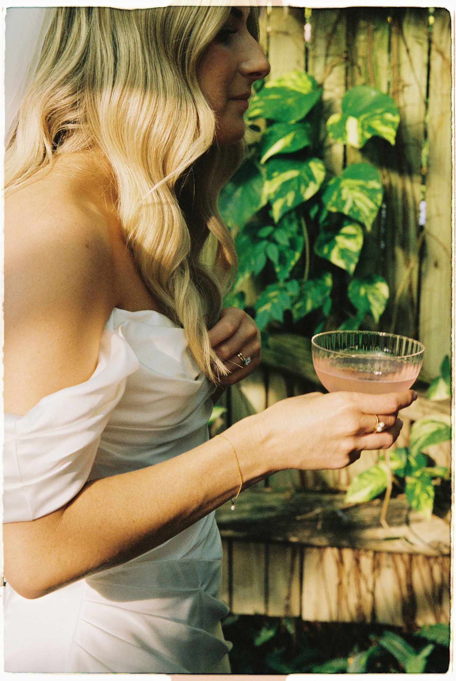 Bride in white off-shoulder gown holding a pink cocktail in a coupe glass amid lush tropical greenery.
