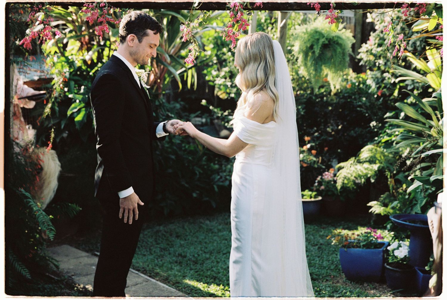 Bride and groom exchange rings in a lush garden wedding ceremony, with the bride in a white gown and groom in a black suit.