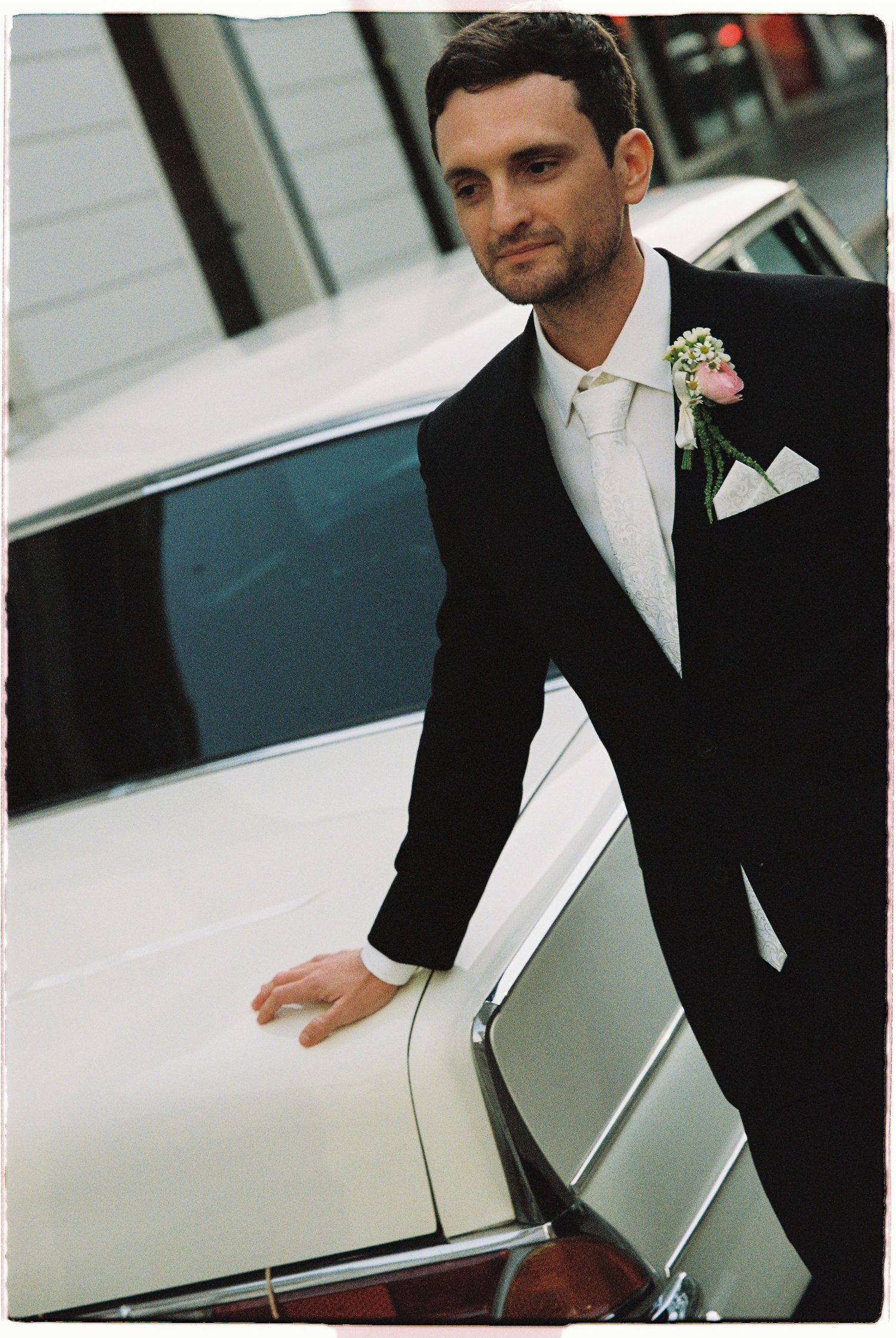 Dapper groom in black tuxedo with pink boutonniere leaning against a vintage white wedding car.