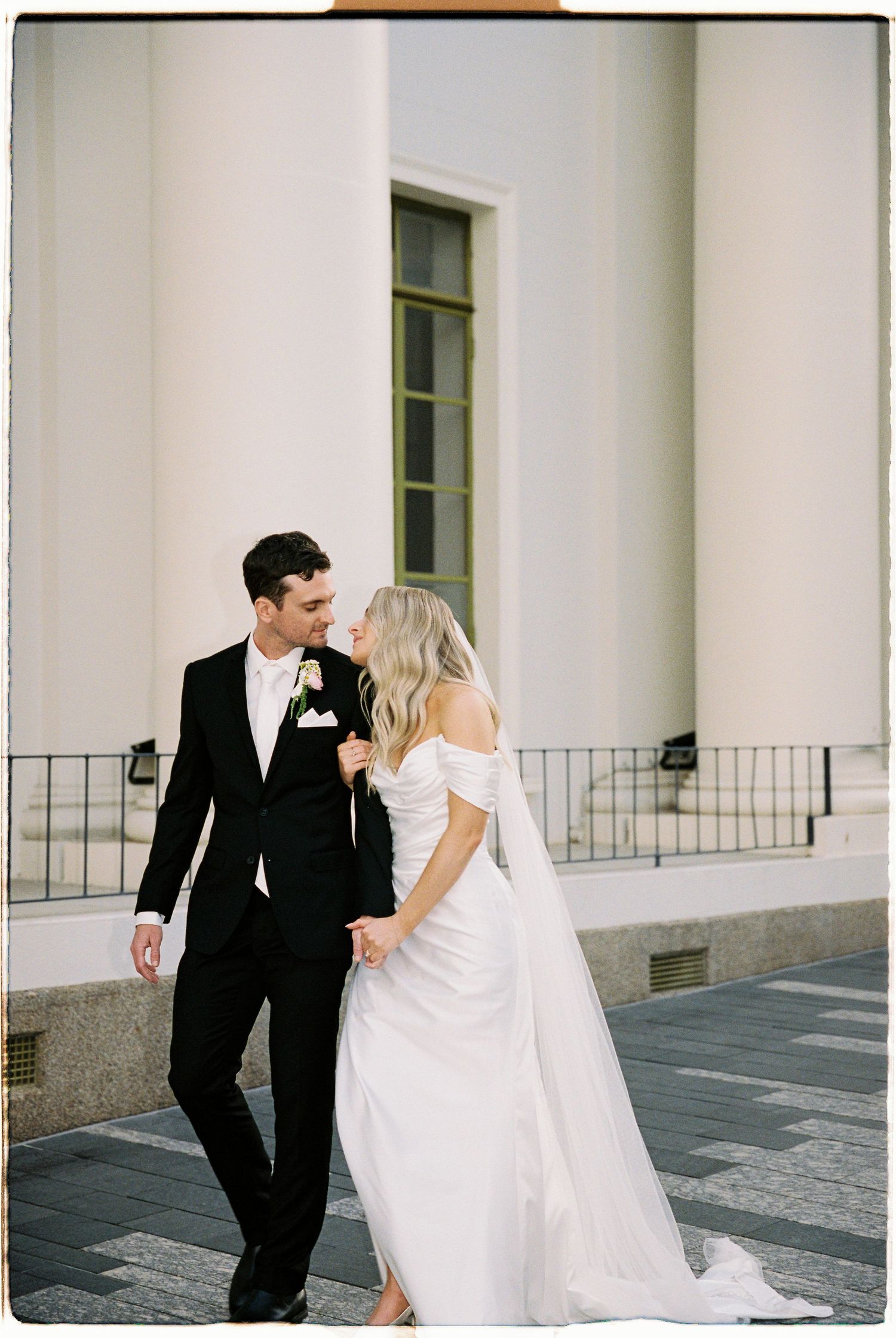 Bride and groom walk hand in hand, sharing a loving gaze beside grand white columns on their wedding day.