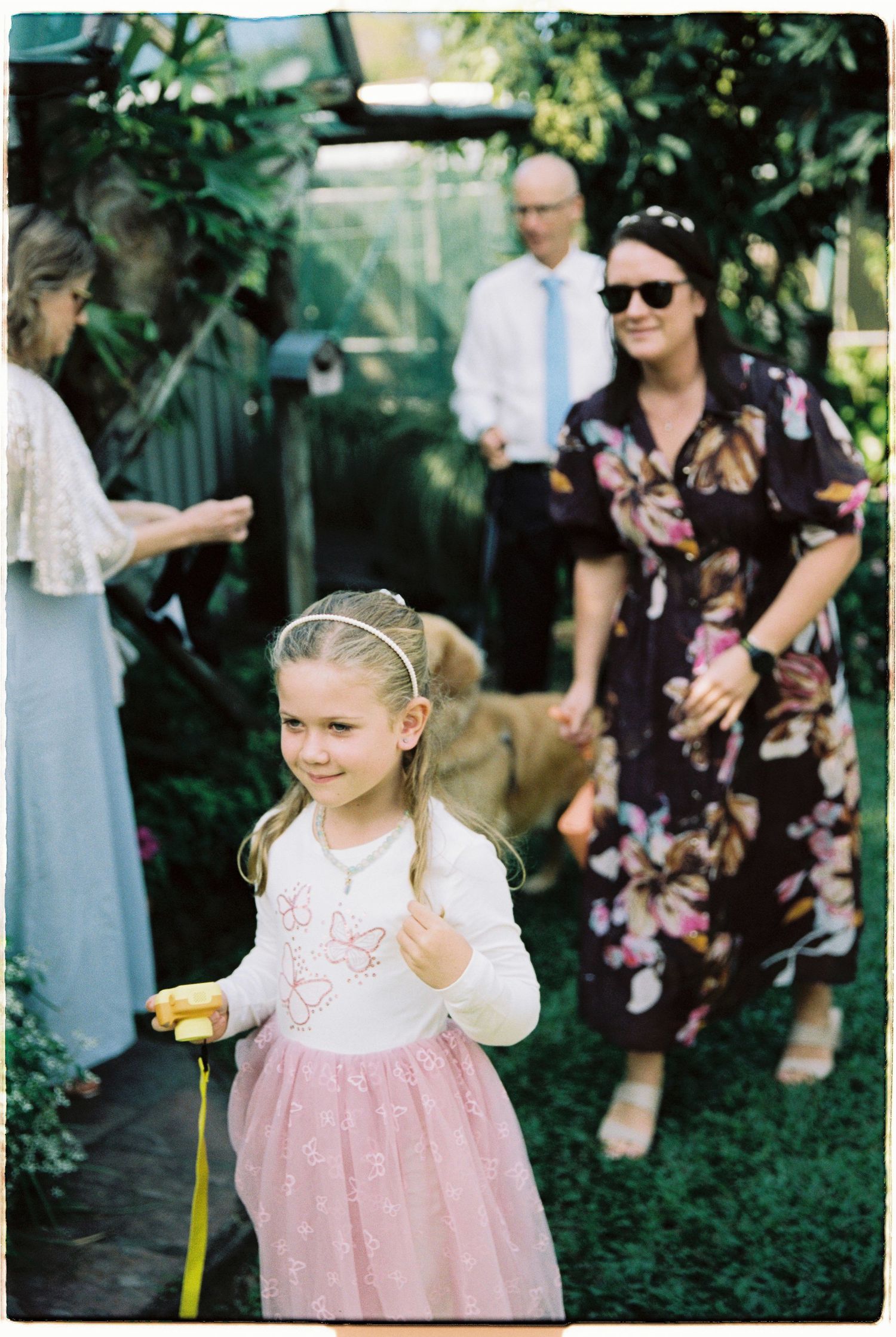 Young flower girl in pink tutu and white top walks through garden during outdoor wedding celebration.