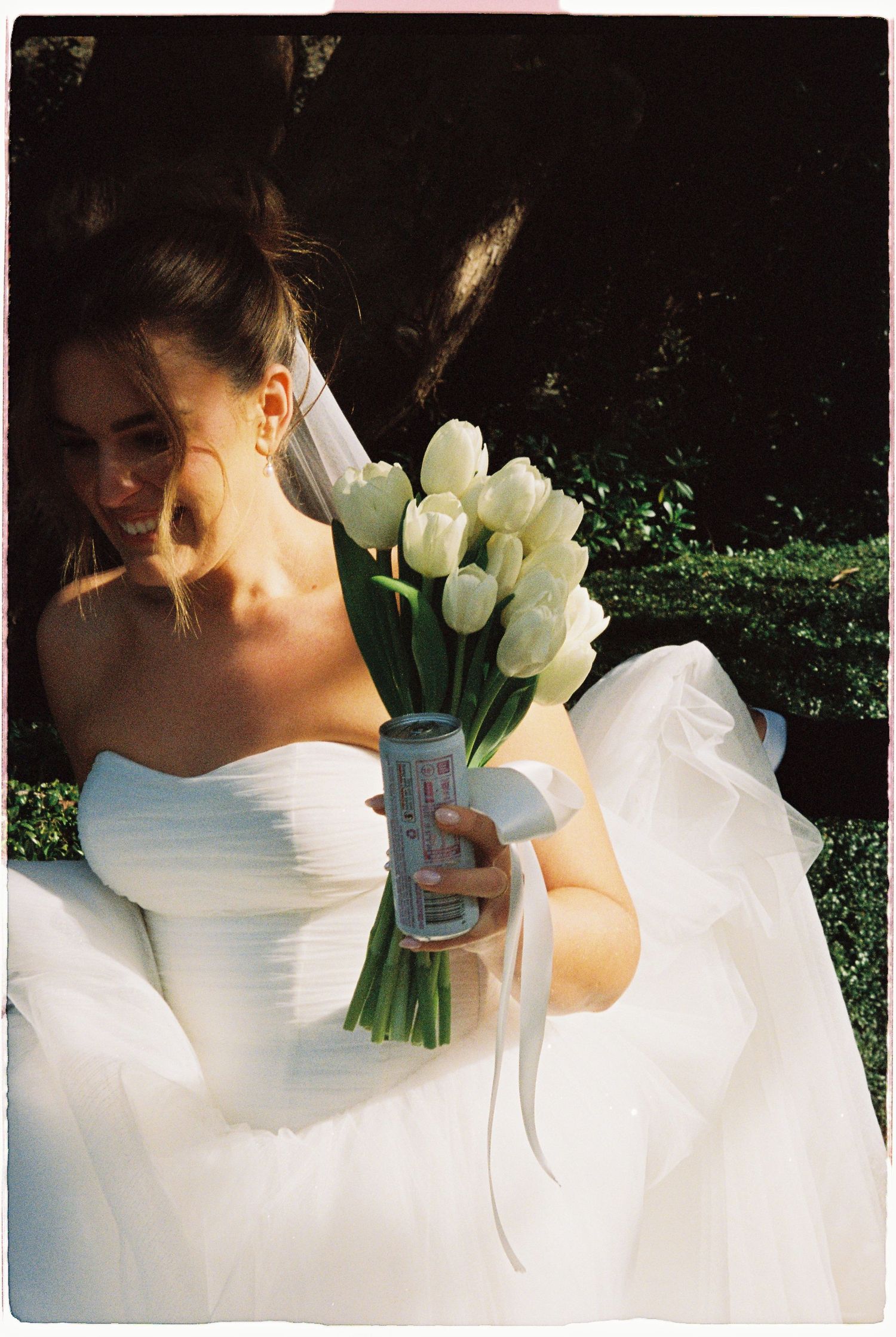 Bride in white strapless gown holding white tulip bouquet and a can outdoors.