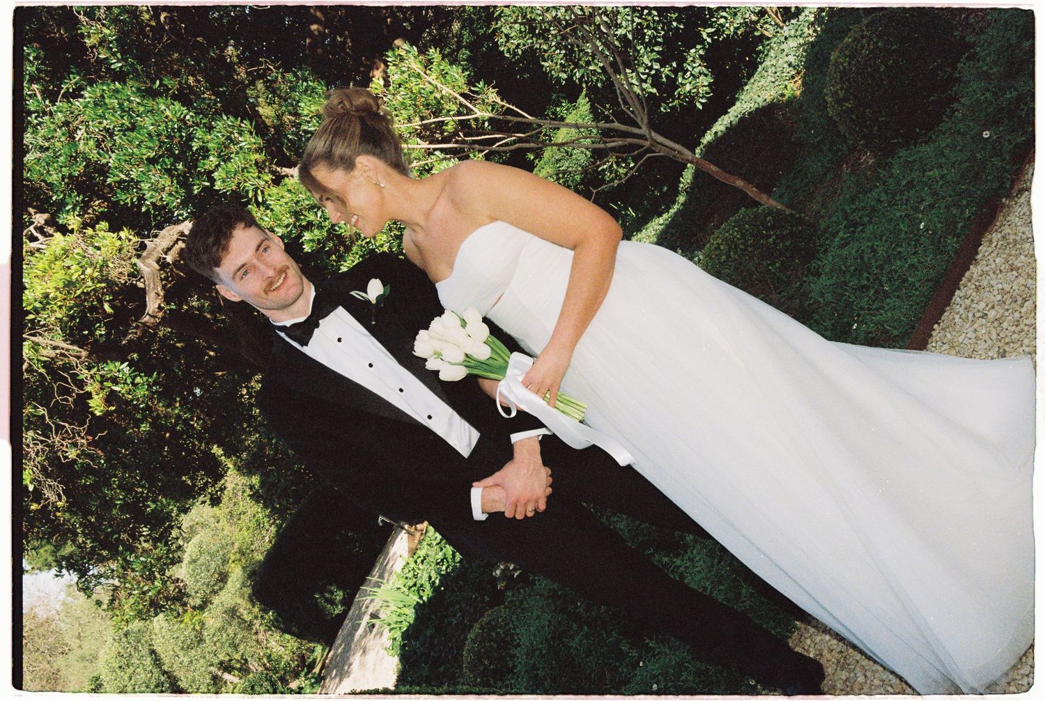 Aerial view of bride in white strapless gown and groom in tuxedo holding hands in lush garden.